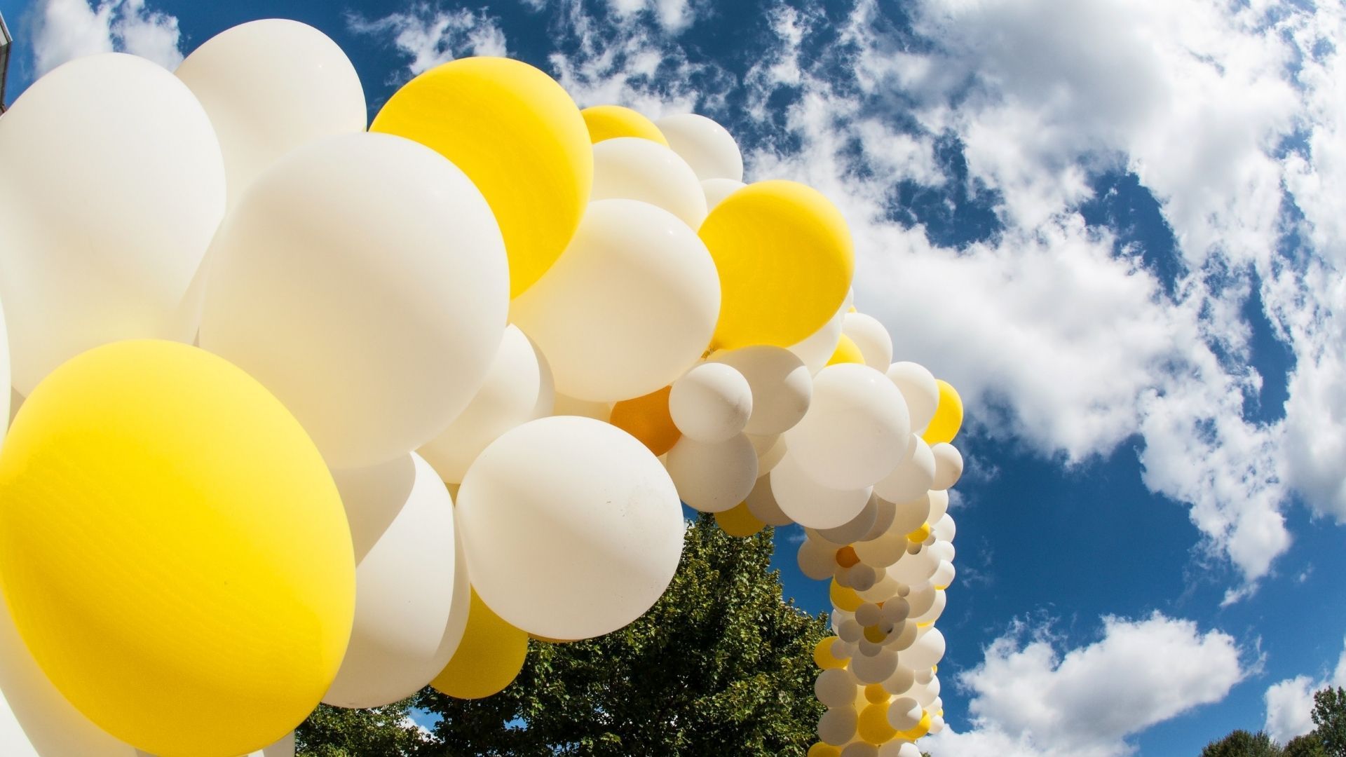 White and yellow balloons forming an arch against a blue sky with clouds