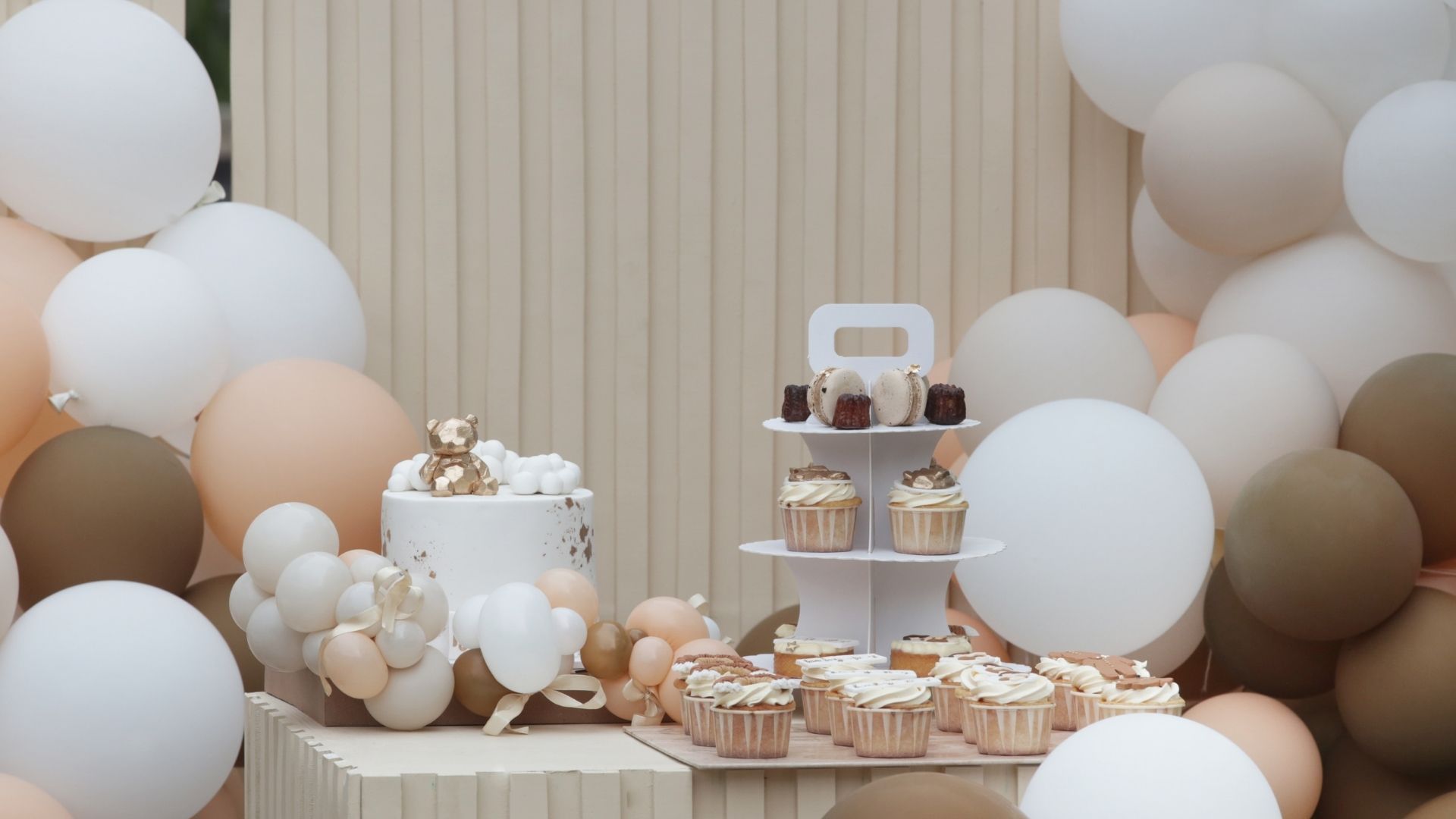 Pastel balloon dessert table with cakes, cupcakes, and treats against a beige striped backdrop