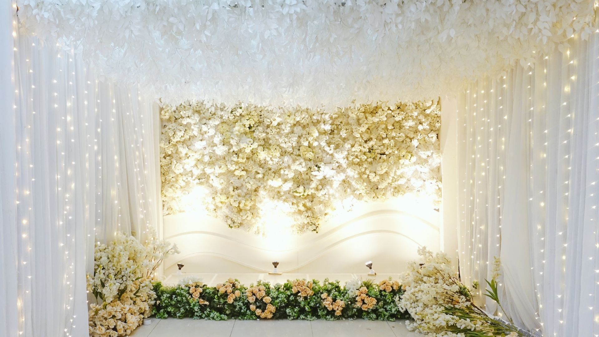 Elegant white and gold floral event backdrop with draped curtains and soft string lights