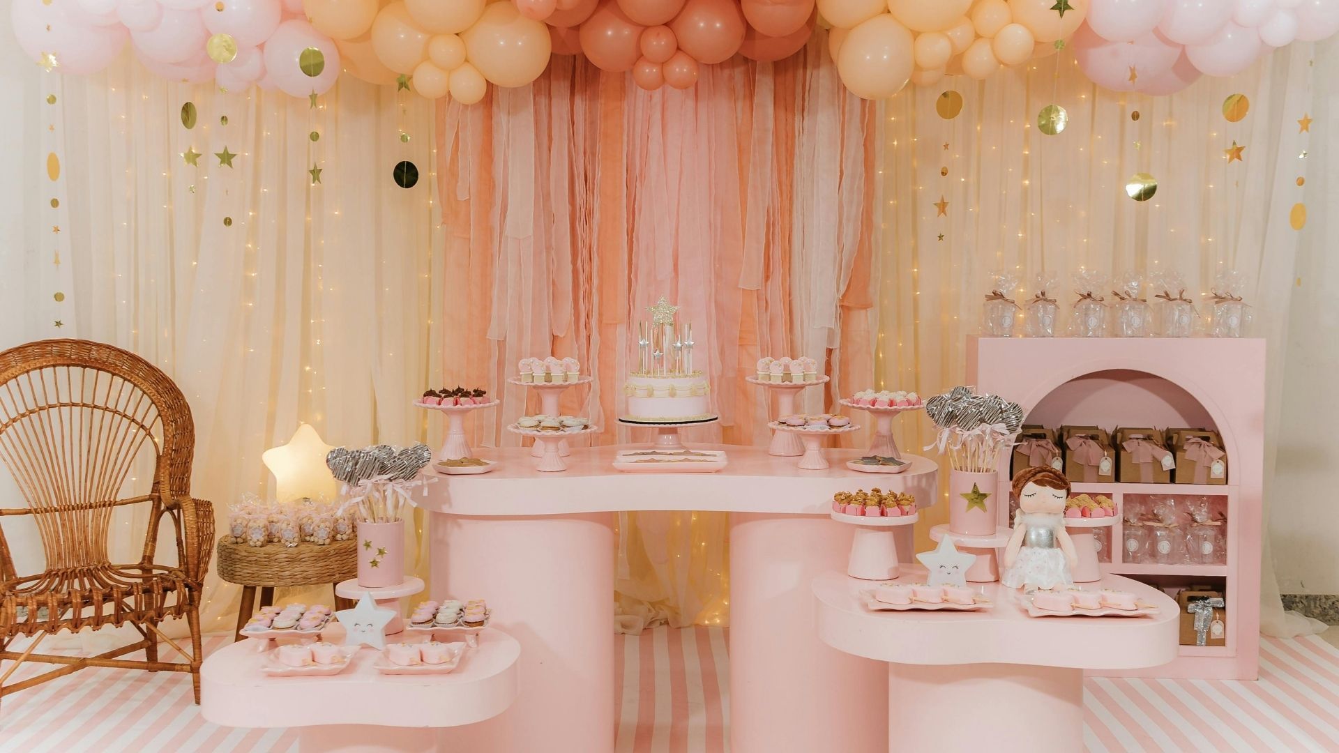 Pastel pink dessert table with balloons, cakes, and tea set at a decorated party backdrop