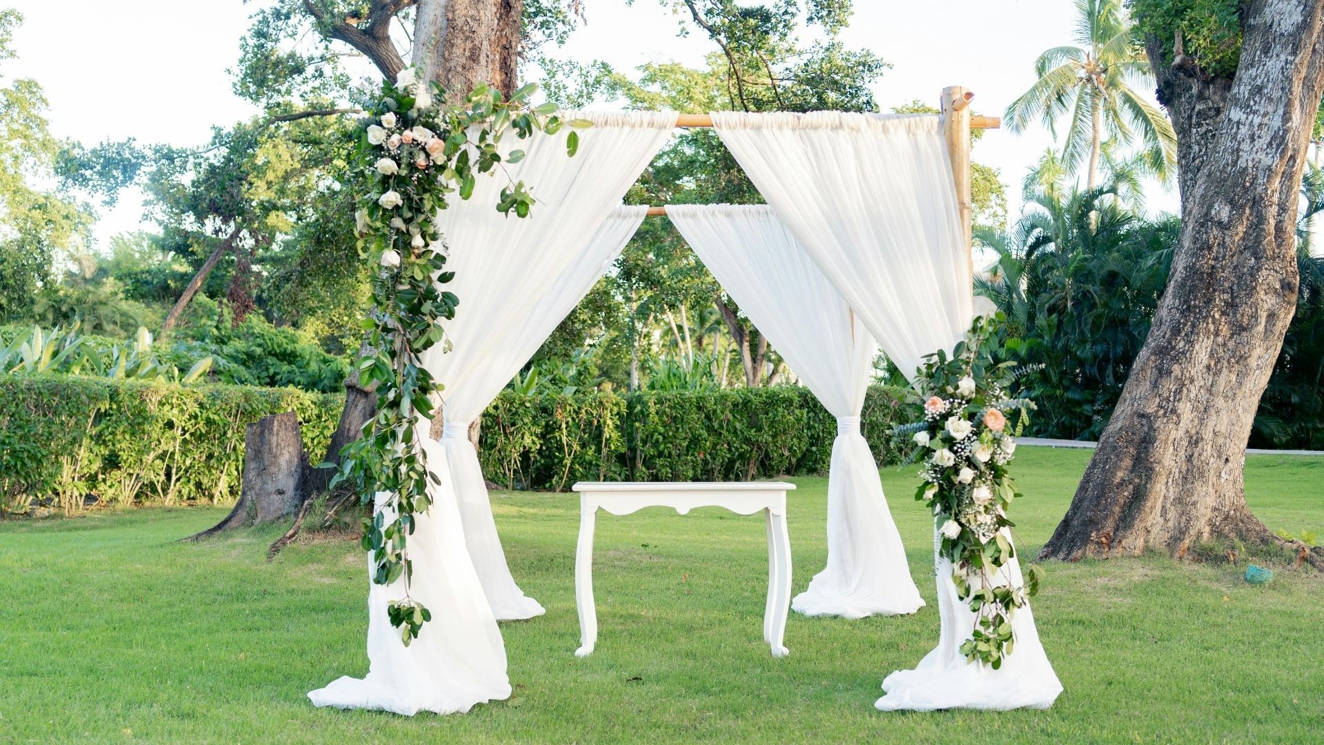 White wedding arch with draped curtains and floral arrangements on a grassy outdoor lawn