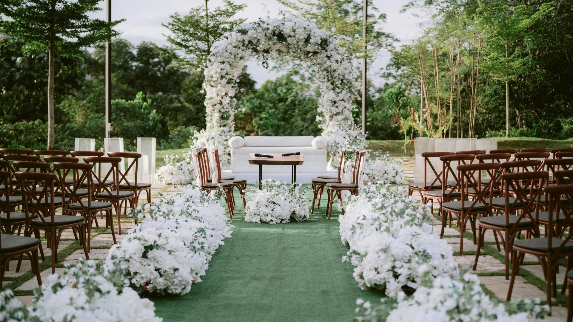 Garden wedding aisle with white floral arch, green carpet, and rows of wooden chairs