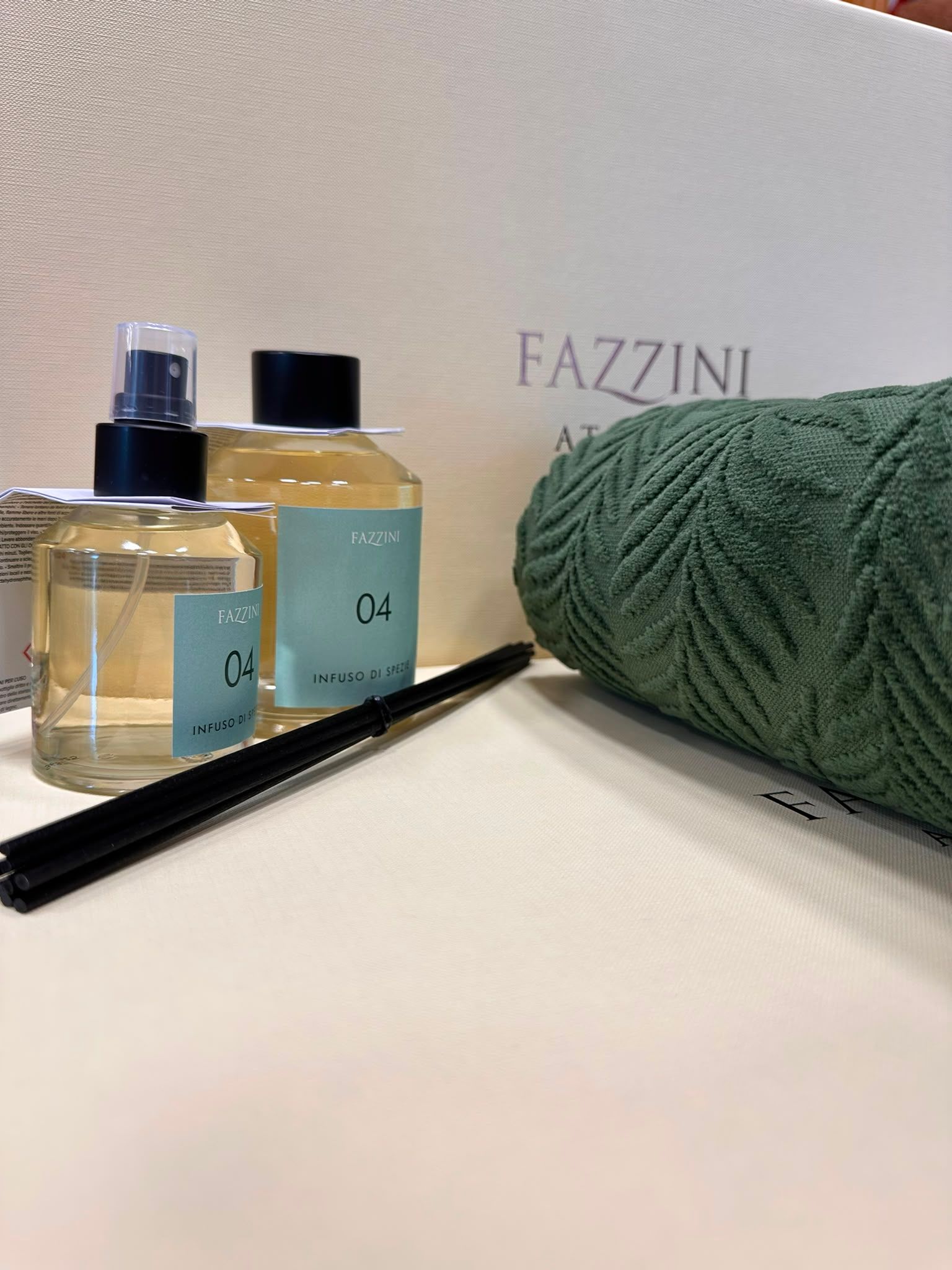 Flaconi di profumo e un pettine nero accanto a un asciugamano verde su un piano di lavoro bianco con il marchio FAZZINI.