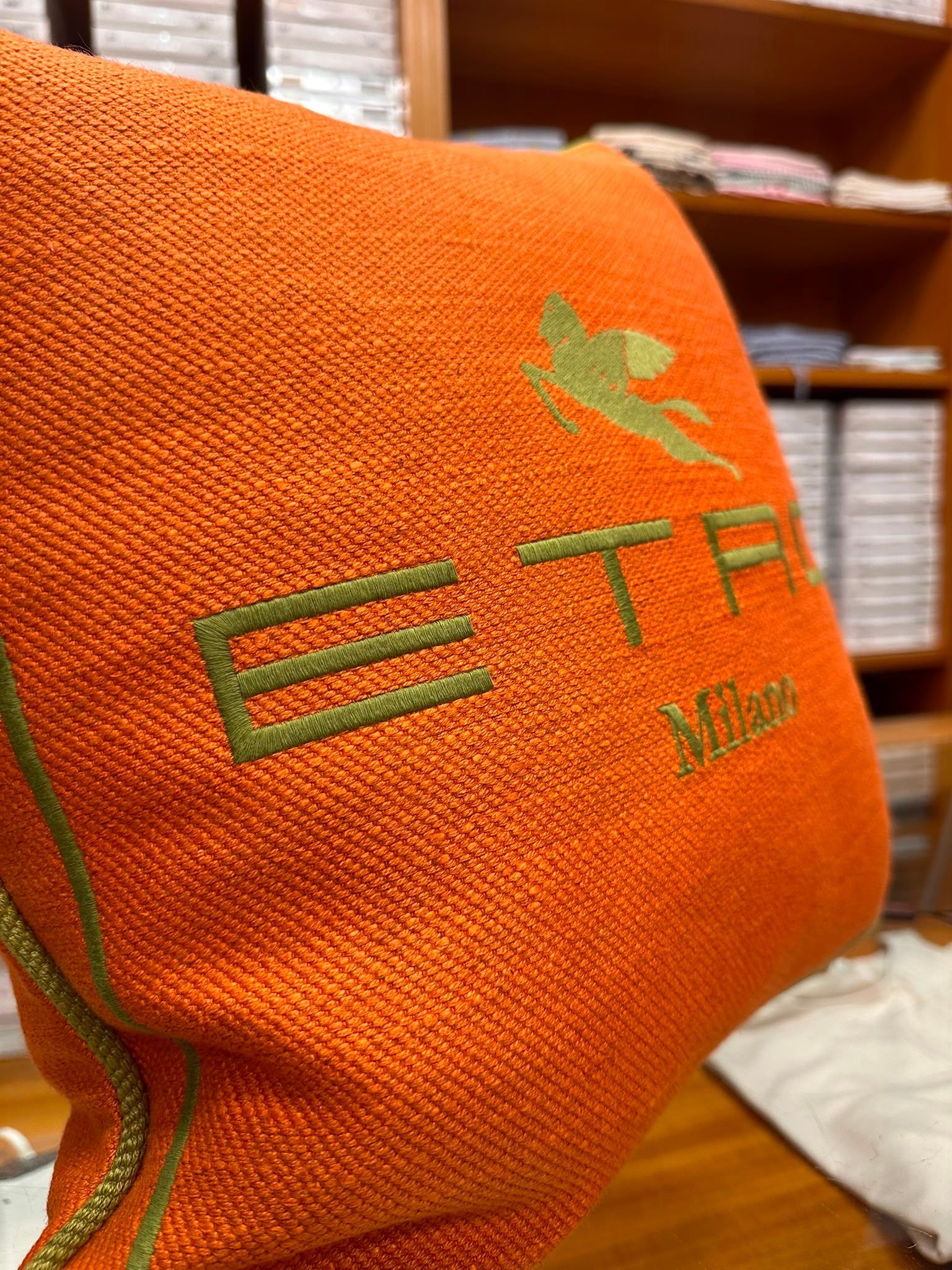 Tessuto arancione ricamato con logo verde, posto davanti a mensole di legno e un panno bianco.