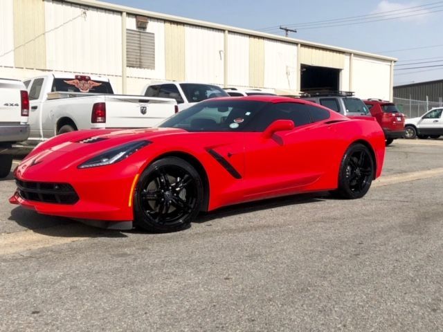 Red sports car parked in an industrial lot beside white trucks and warehouse buildings.
