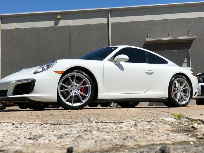 White Porsche coupe parked outside a gray building, side view on gravel.