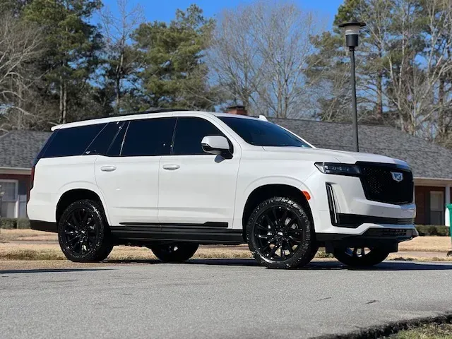 White SUV with black wheels parked on a paved road beside trees under a blue sky