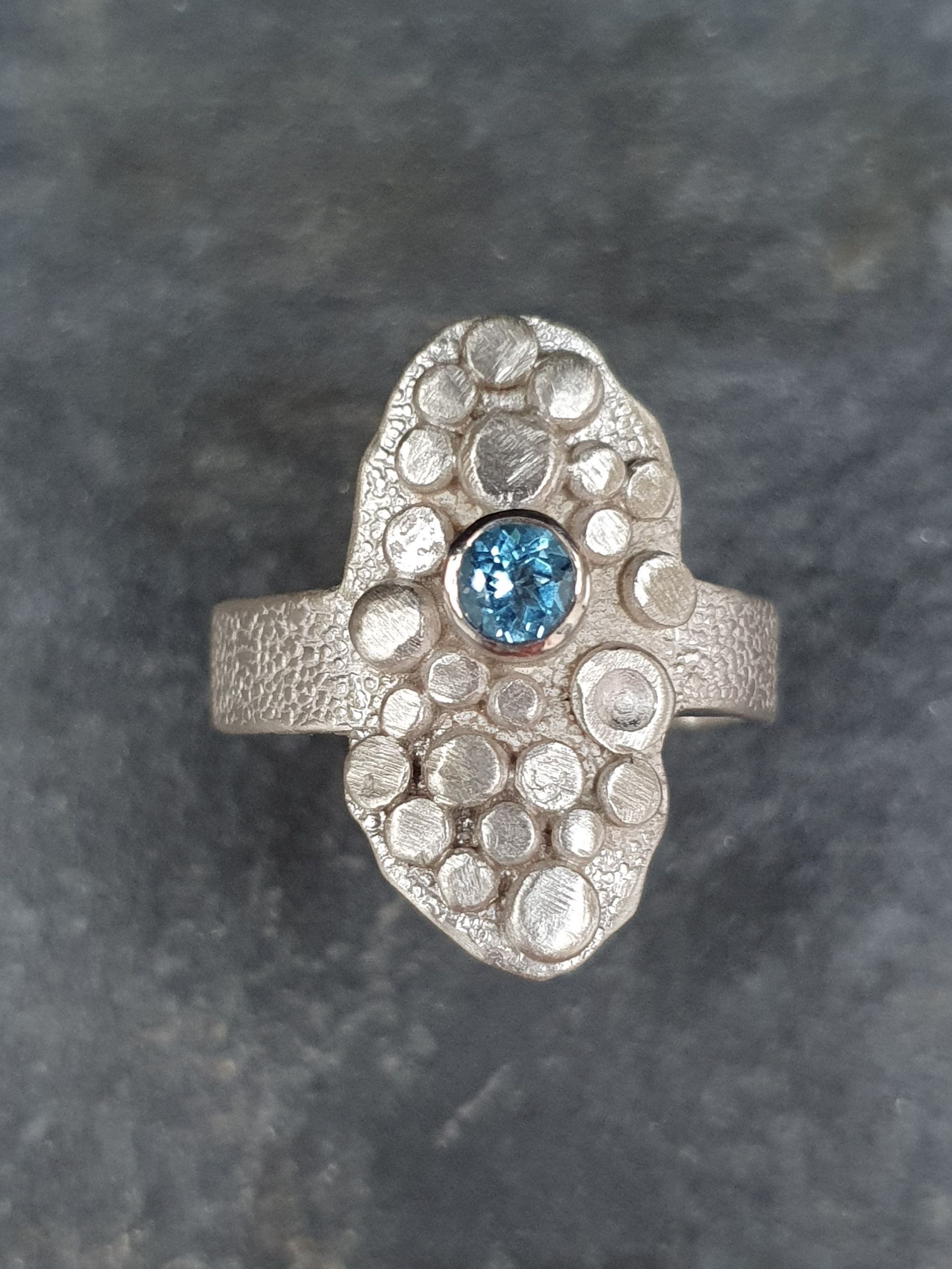 Zilveren ring met een gestructureerde band, ovale bovenkant met diverse verhoogde zilveren cirkels en een blauwe topaas.