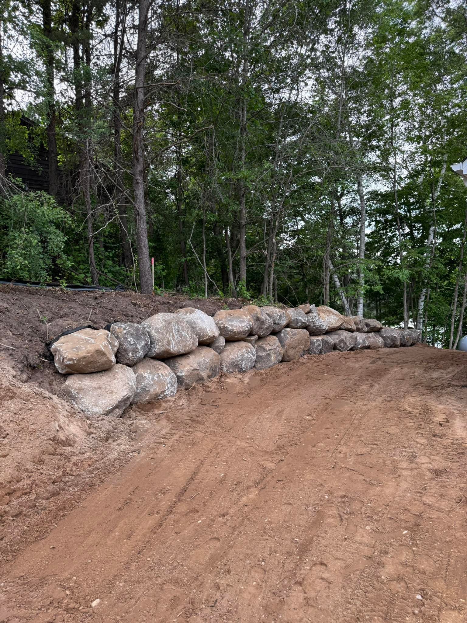 A low retaining wall made of stacked natural rocks separates a freshly graded dirt slope from a dense forest background.