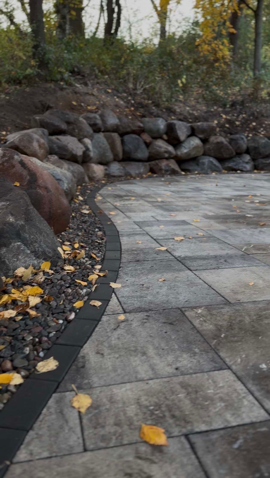 A stone patio with a curved dark border, set against a stone retaining wall and a wooded area with scattered autumn leaves.