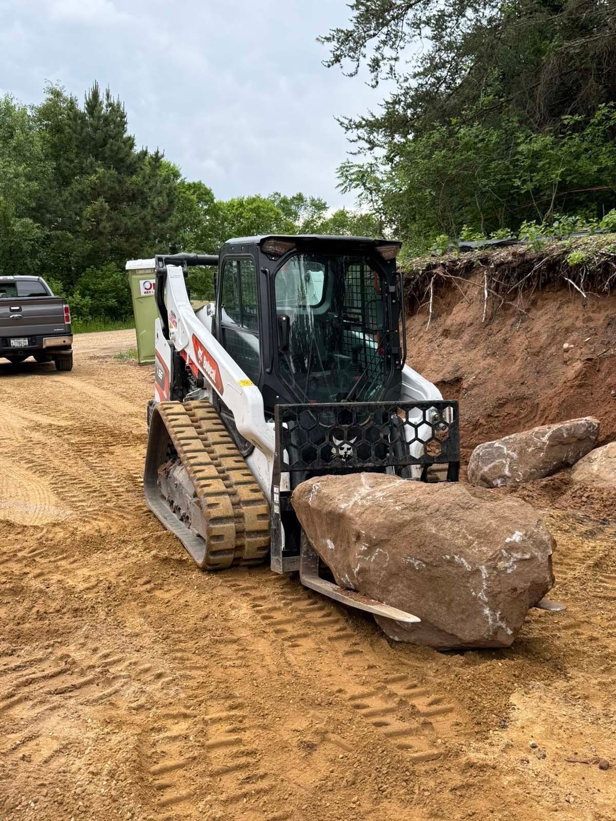 A white tracked skid steer loader uses a grapple attachment to lift a large boulder at a construction site.
