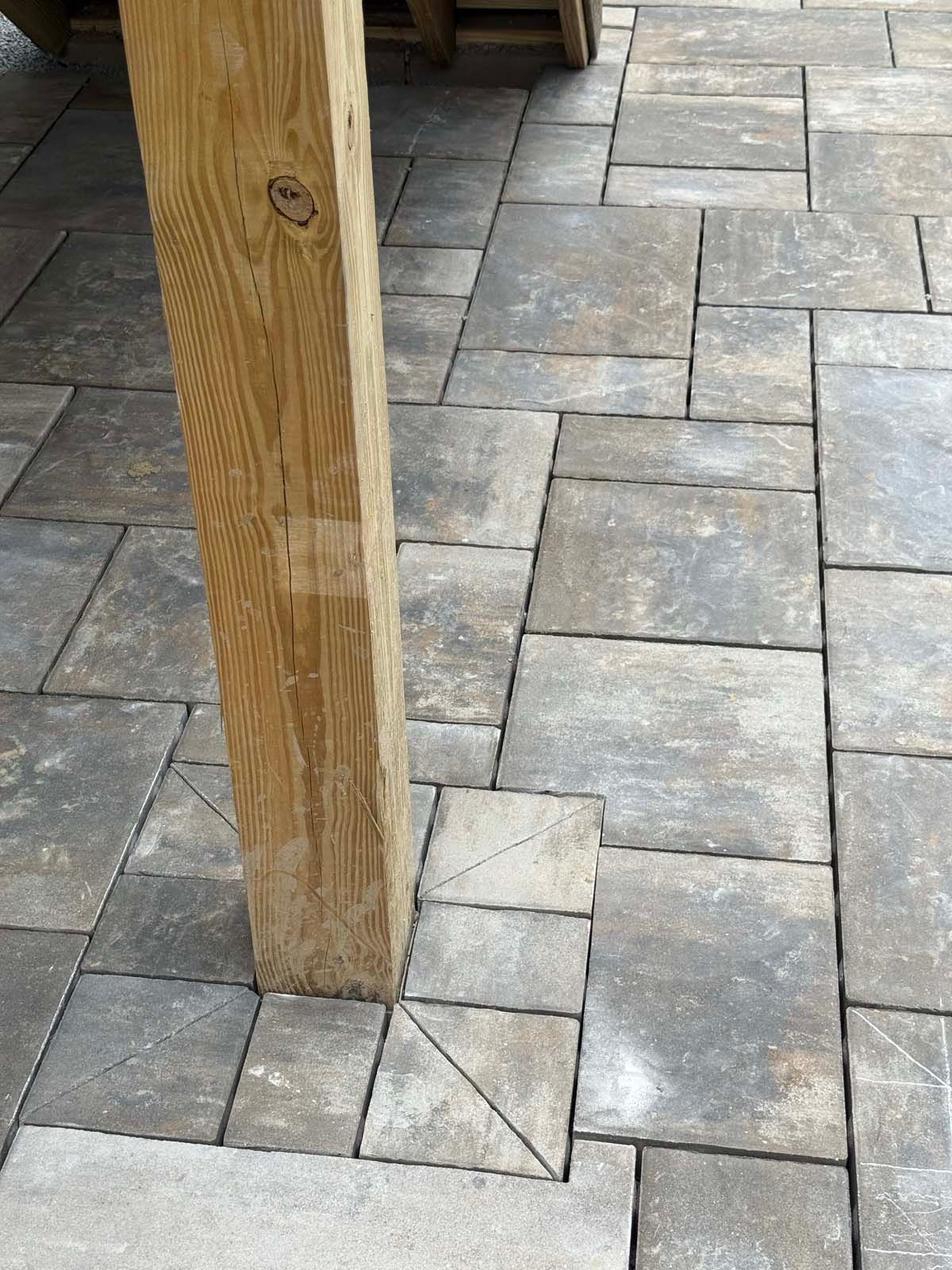 A wooden post stands on a paved patio made of rectangular, multi-toned gray stone tiles fitted precisely around its base.