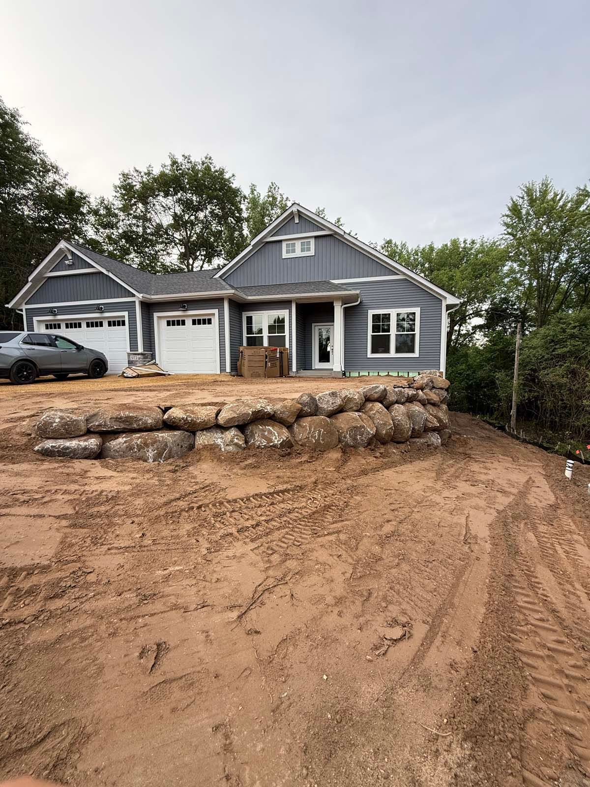A blue suburban house under construction with a newly built stone retaining wall and tire tracks on a sandy lot.
