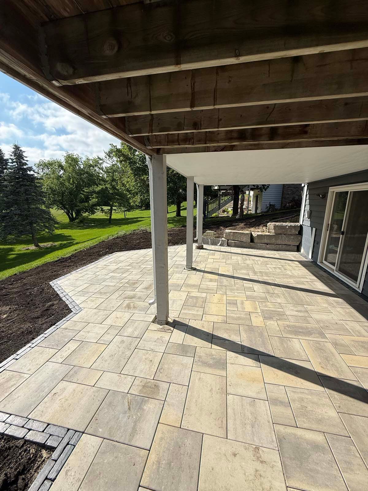 A paved patio with tan and grey rectangular stones sits under a wooden deck with a view of a grassy lawn and trees.