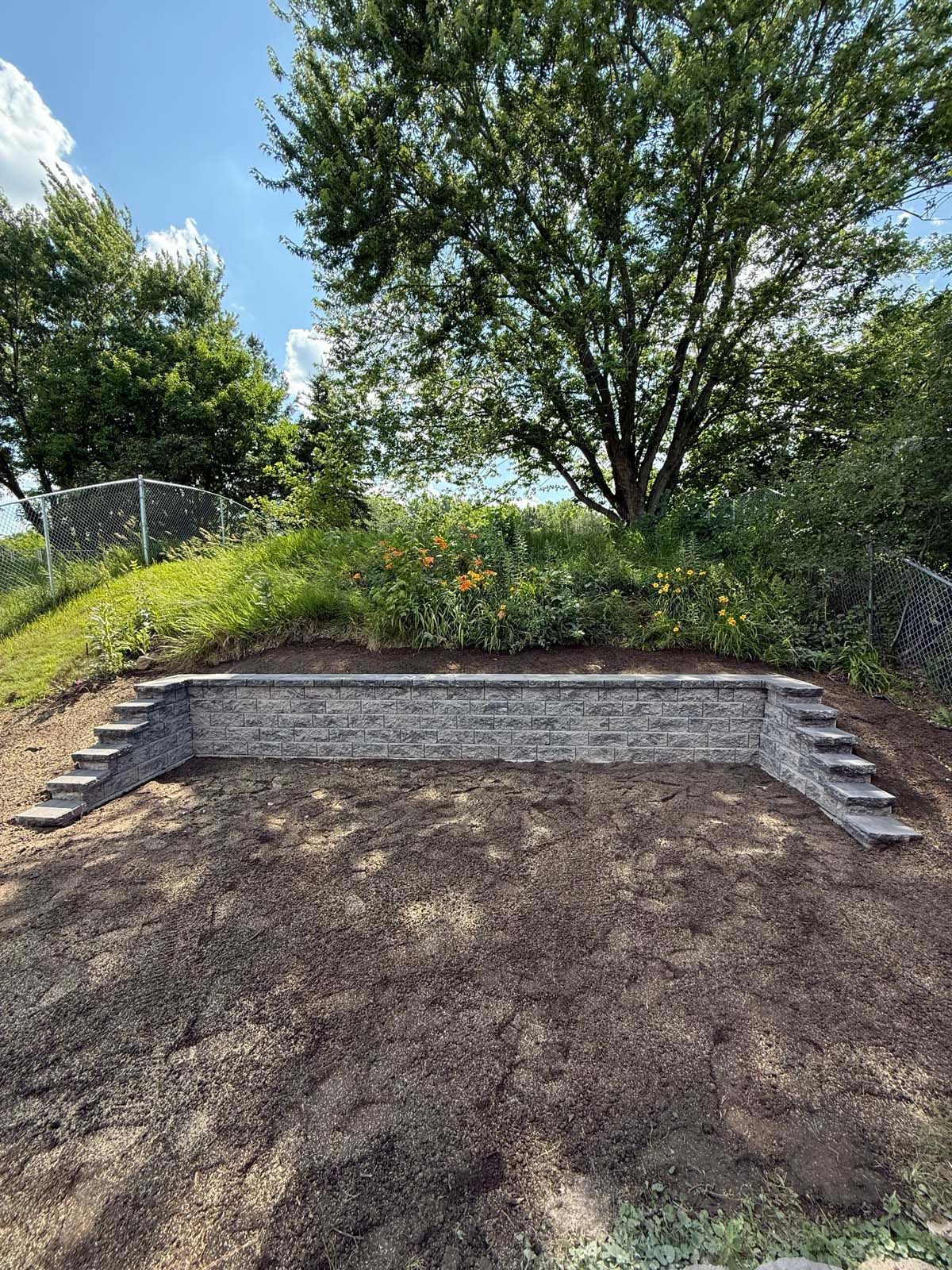 A newly built gray stone retaining wall with matching steps on both sides, situated at the base of a grassy hill.