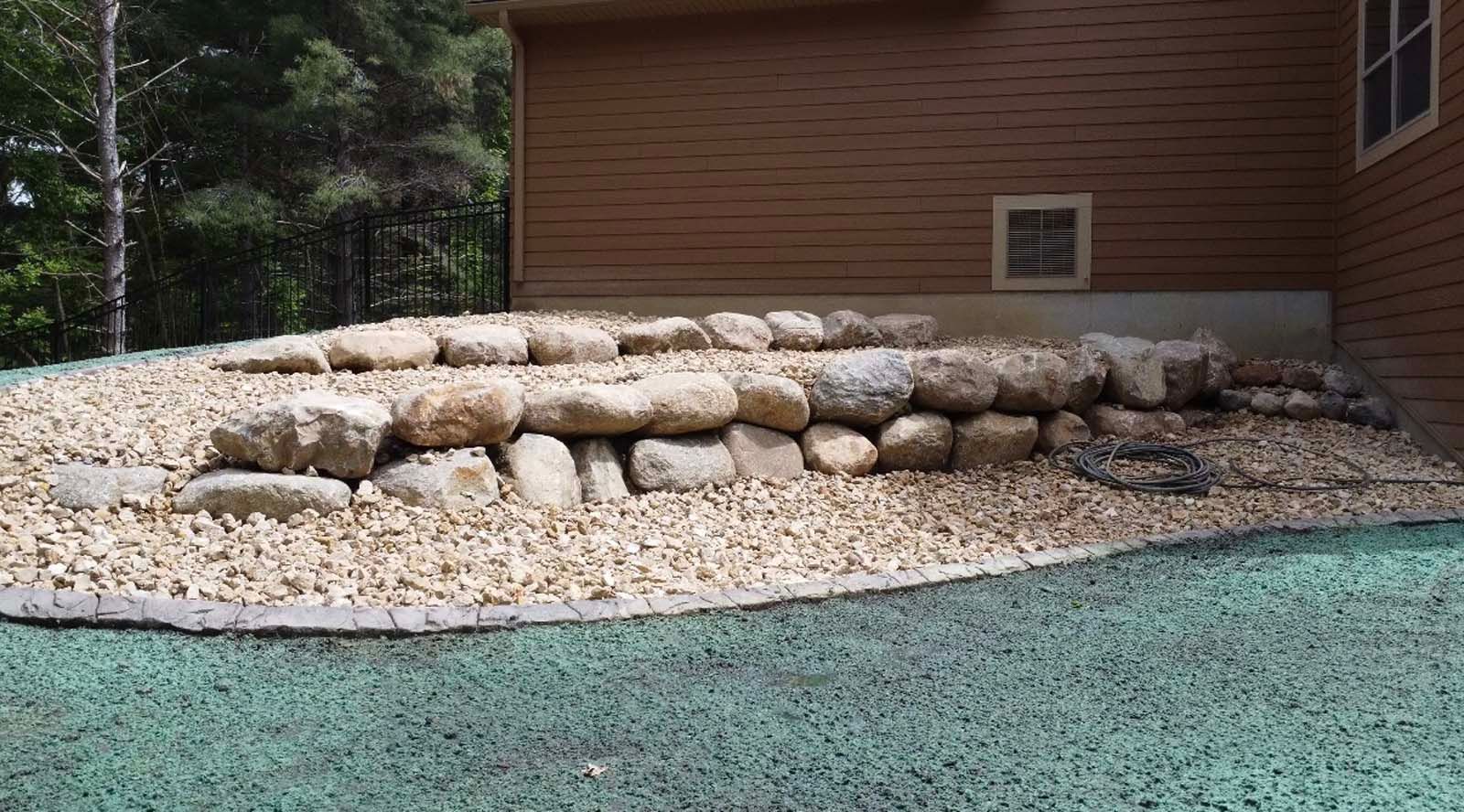 A multi-tiered stone retaining wall set against the side of a brown house, surrounded by light-colored gravel.