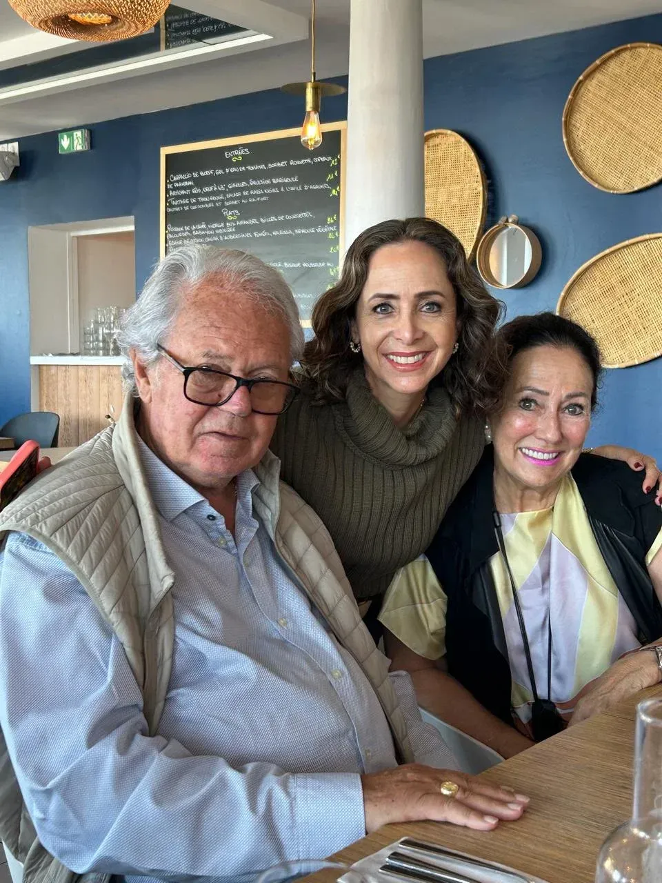 Tres personas sonriendo en una mesa de café, con una decoración de pared tejida y una pizarra con el menú detrás de ellas.