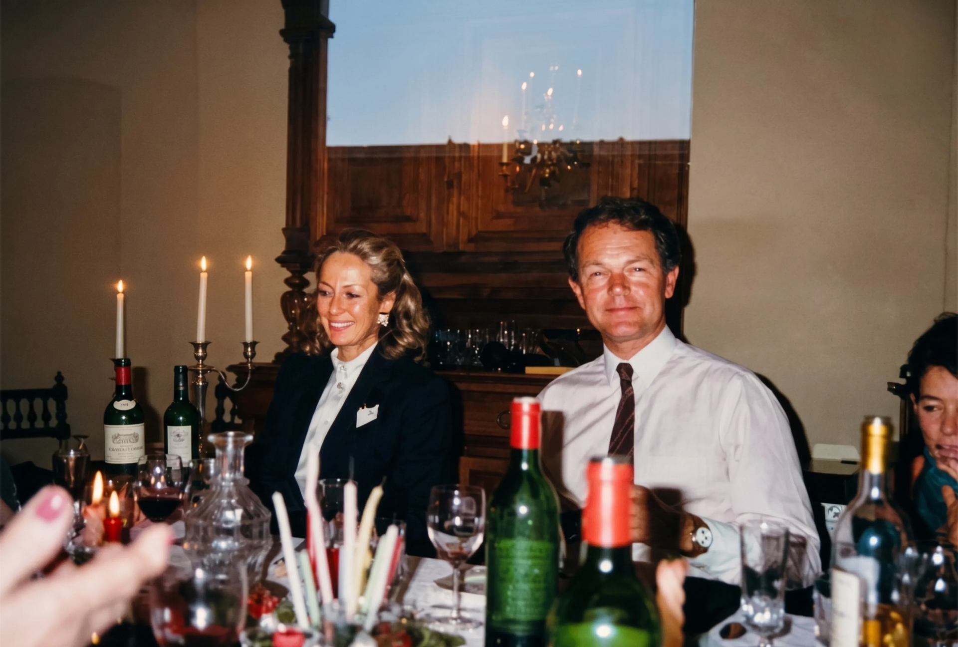 Personas sentadas a una mesa iluminada con velas, con botellas y copas de vino, en un comedor formal.