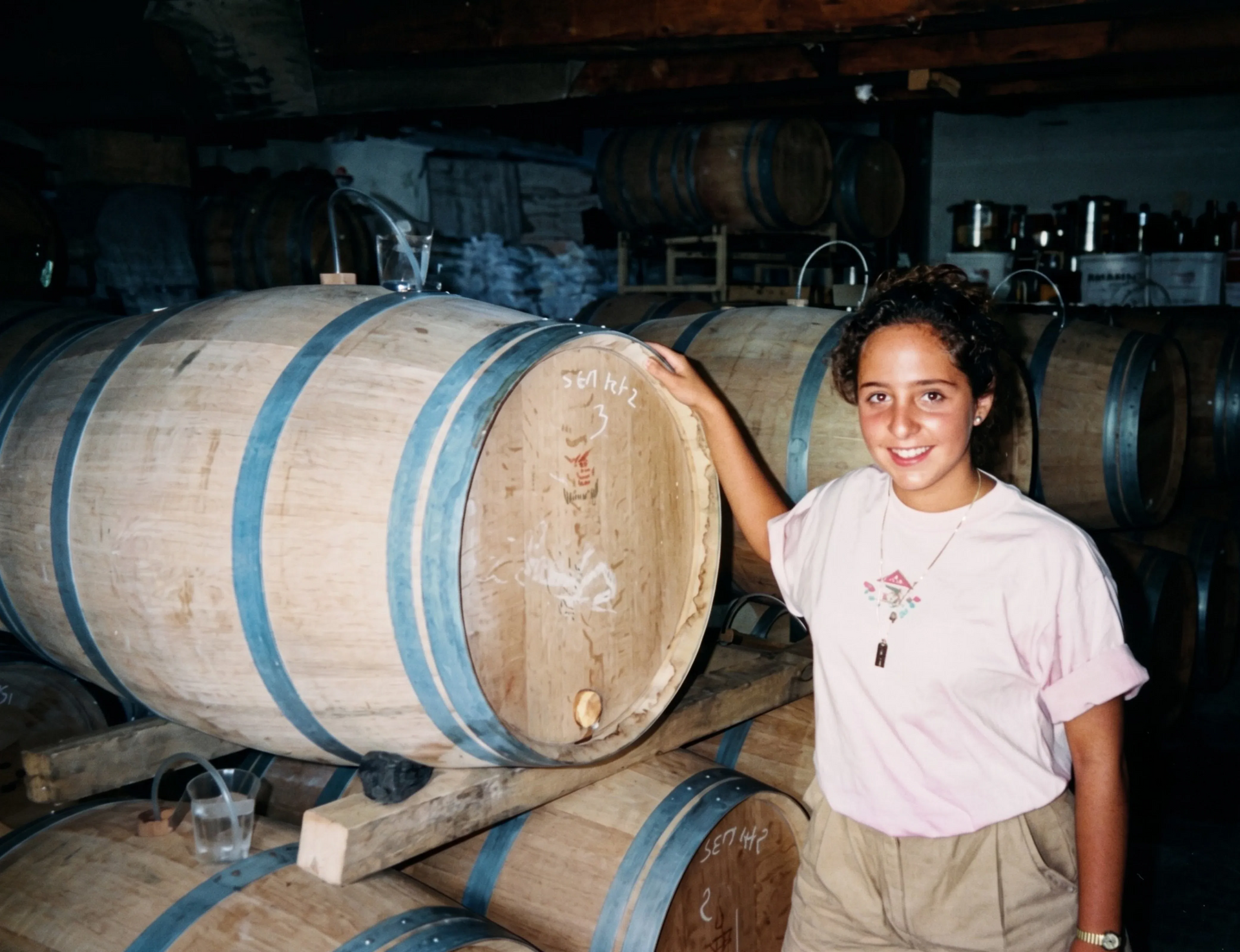 Una persona sonríe junto a una pila de barriles de madera en una bodega de vinos con poca luz.