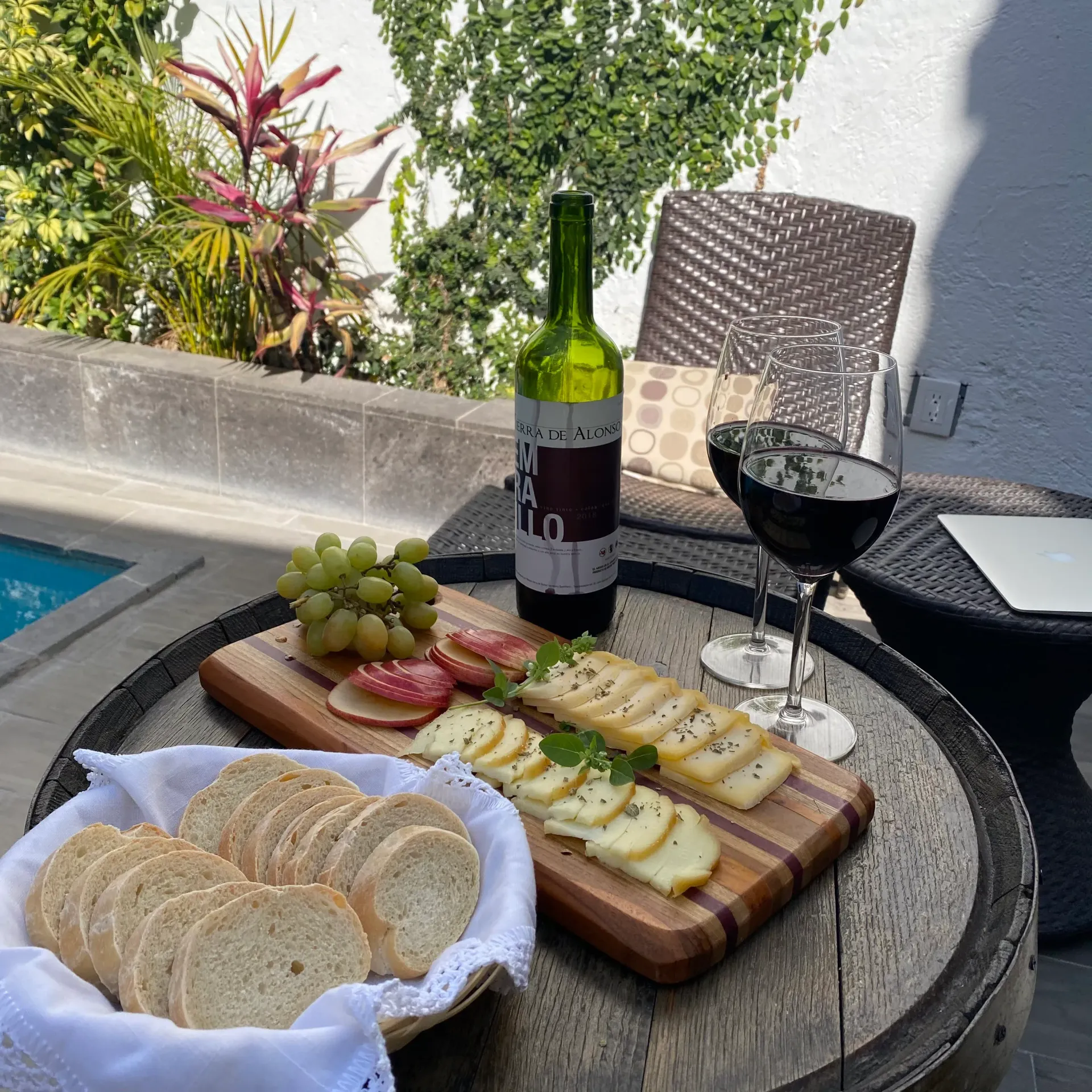 Mesa de patio exterior con vino, queso, galletas saladas, uvas y una copa de cóctel negra junto a la piscina.