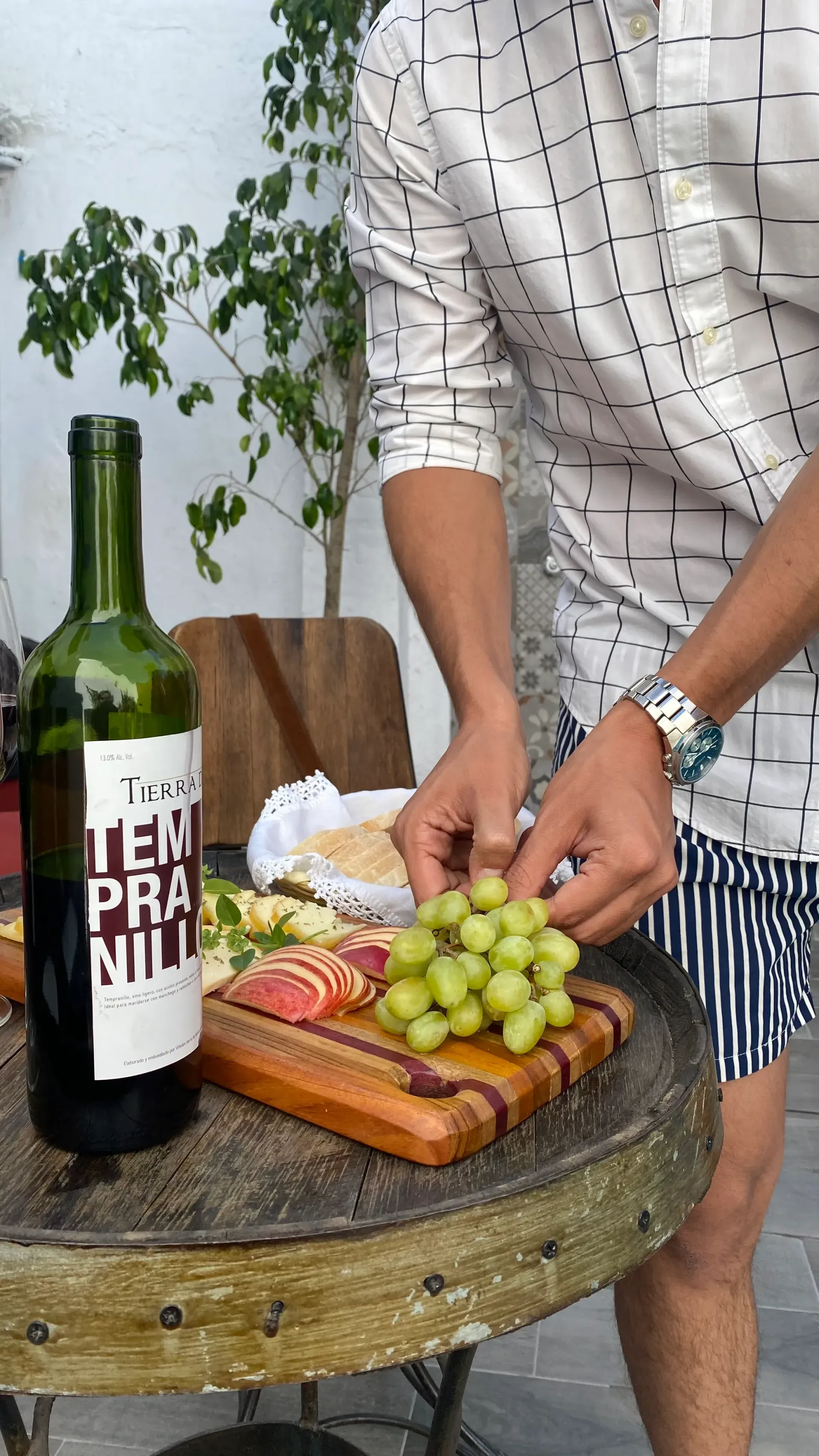 Person arranging grapes and snacks on a small round table beside a wine bottle outdoors