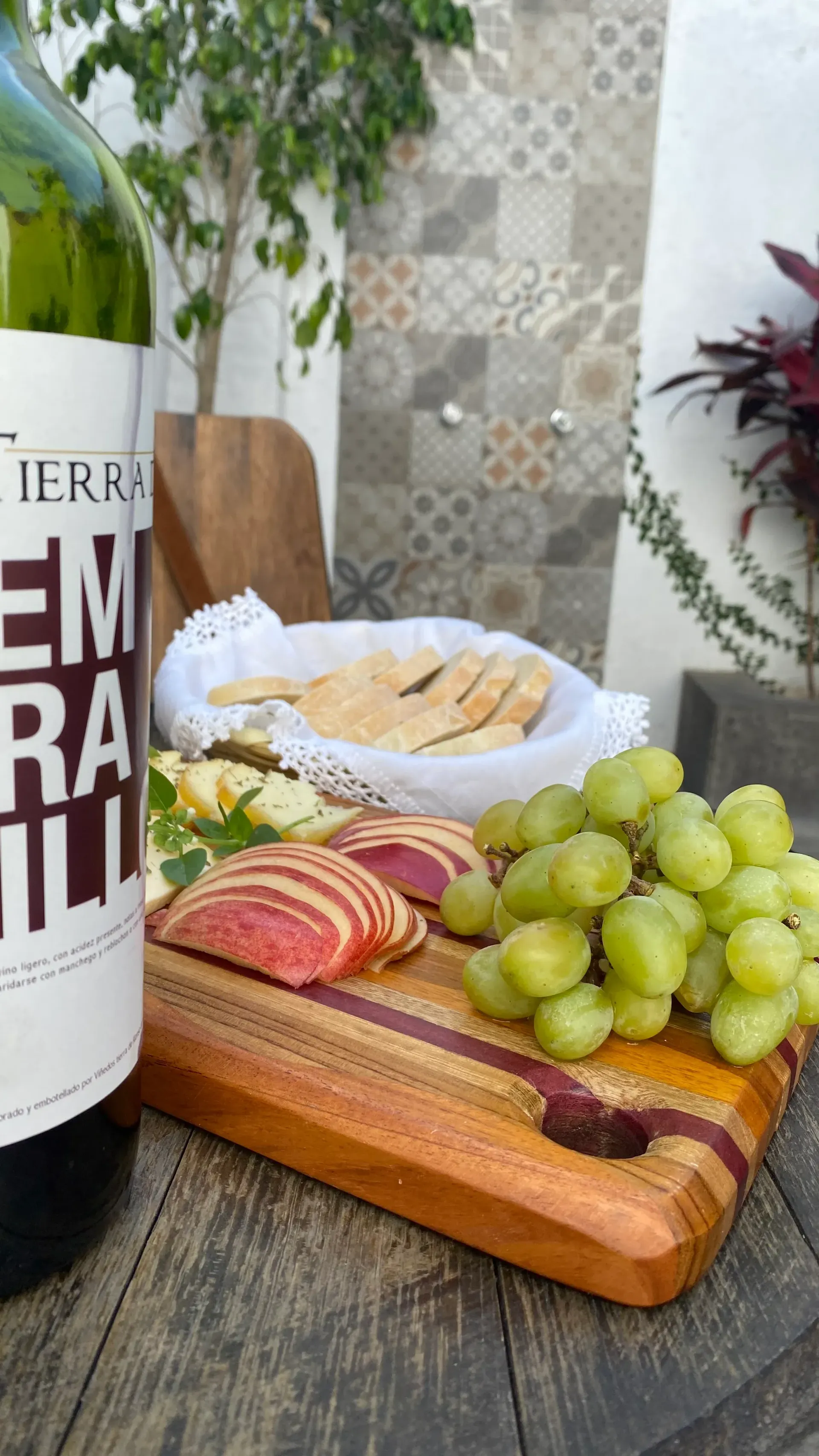 Queso y fruta en rodajas sobre una tabla de madera junto a una botella de vino en una mesa rústica.