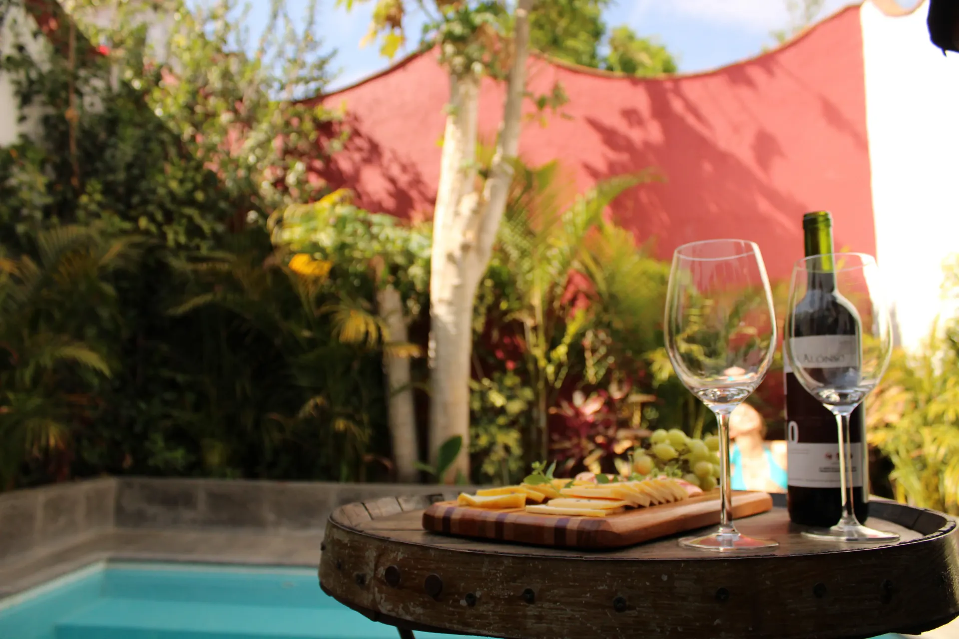 Vino y queso en una mesa de patio junto a la piscina, con plantas tropicales y una vela de sombra rosa.