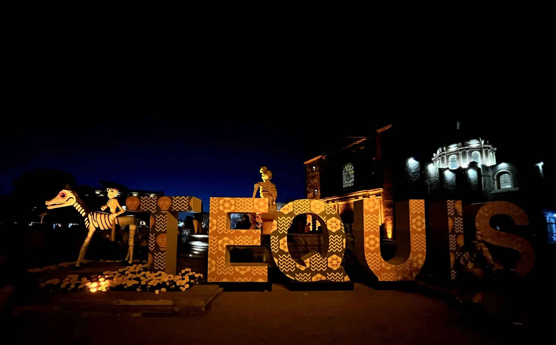 Nighttime light display spelling “EQUIS” in gold, with festive decorations and a lit building in the background