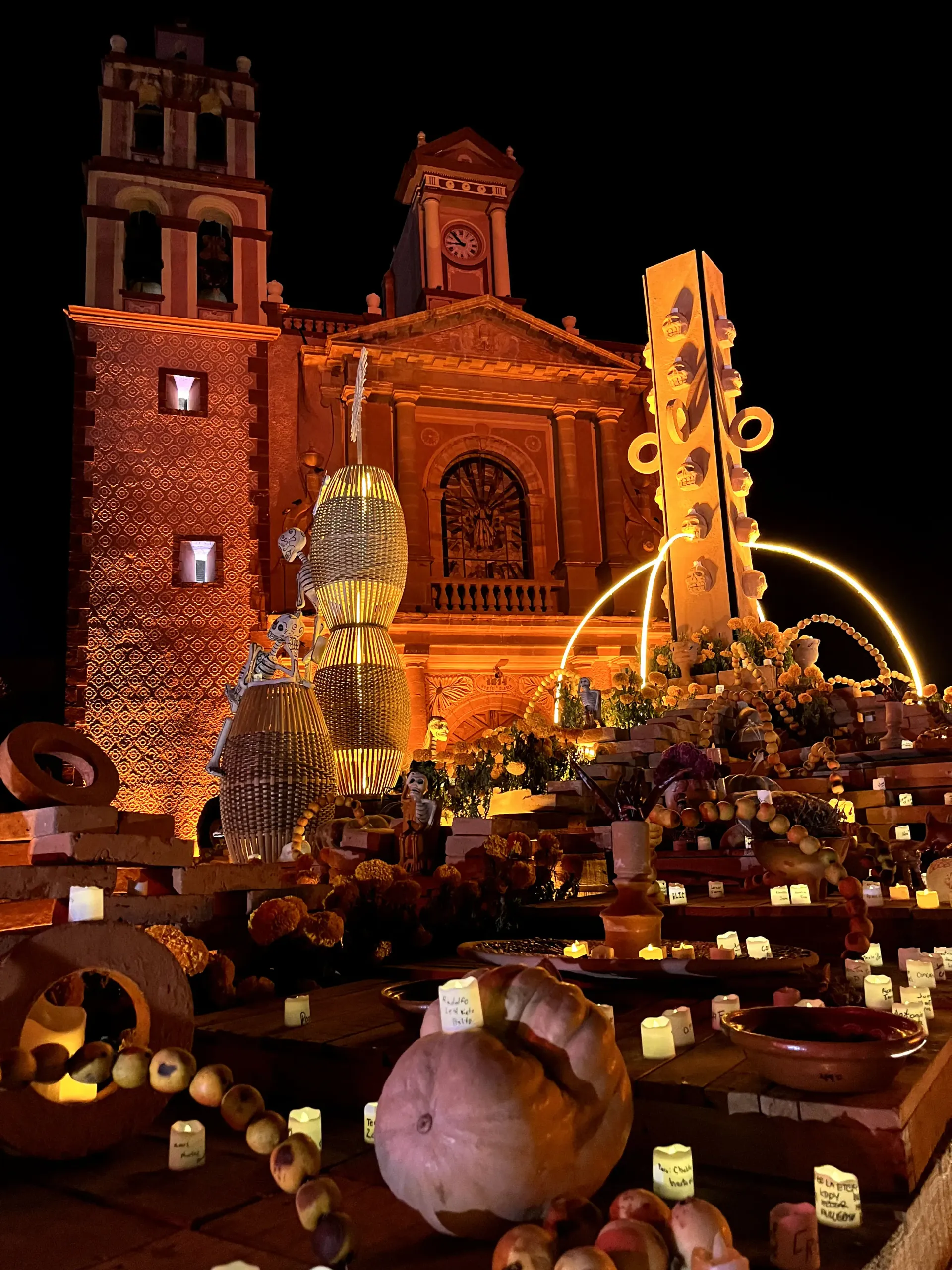 Nighttime church plaza with glowing lights, decorated pumpkins, and a tall illuminated structure
