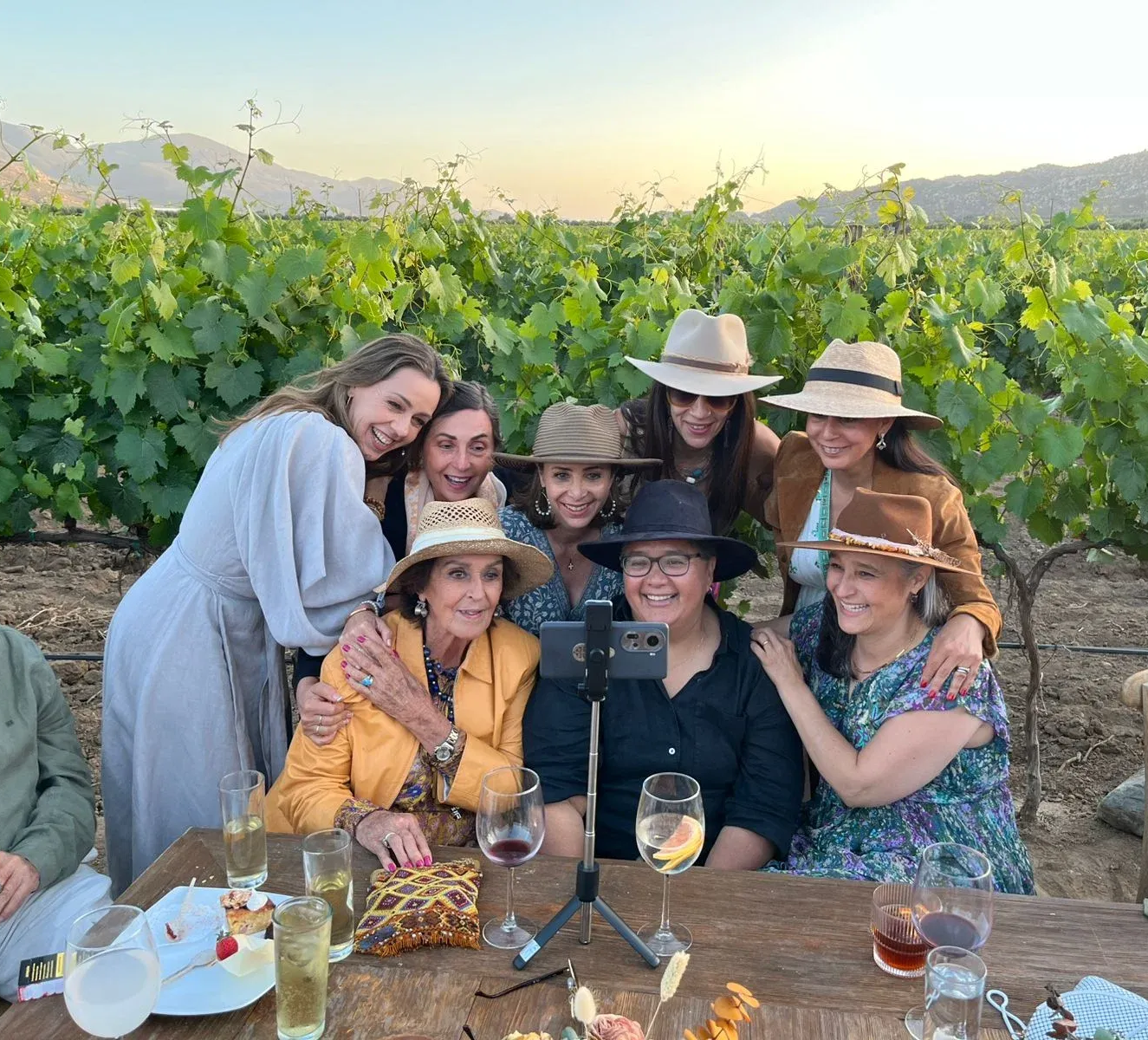 Grupo de amigas con sombreros y vestidos posando en una mesa de viñedo con bebidas al atardecer.
