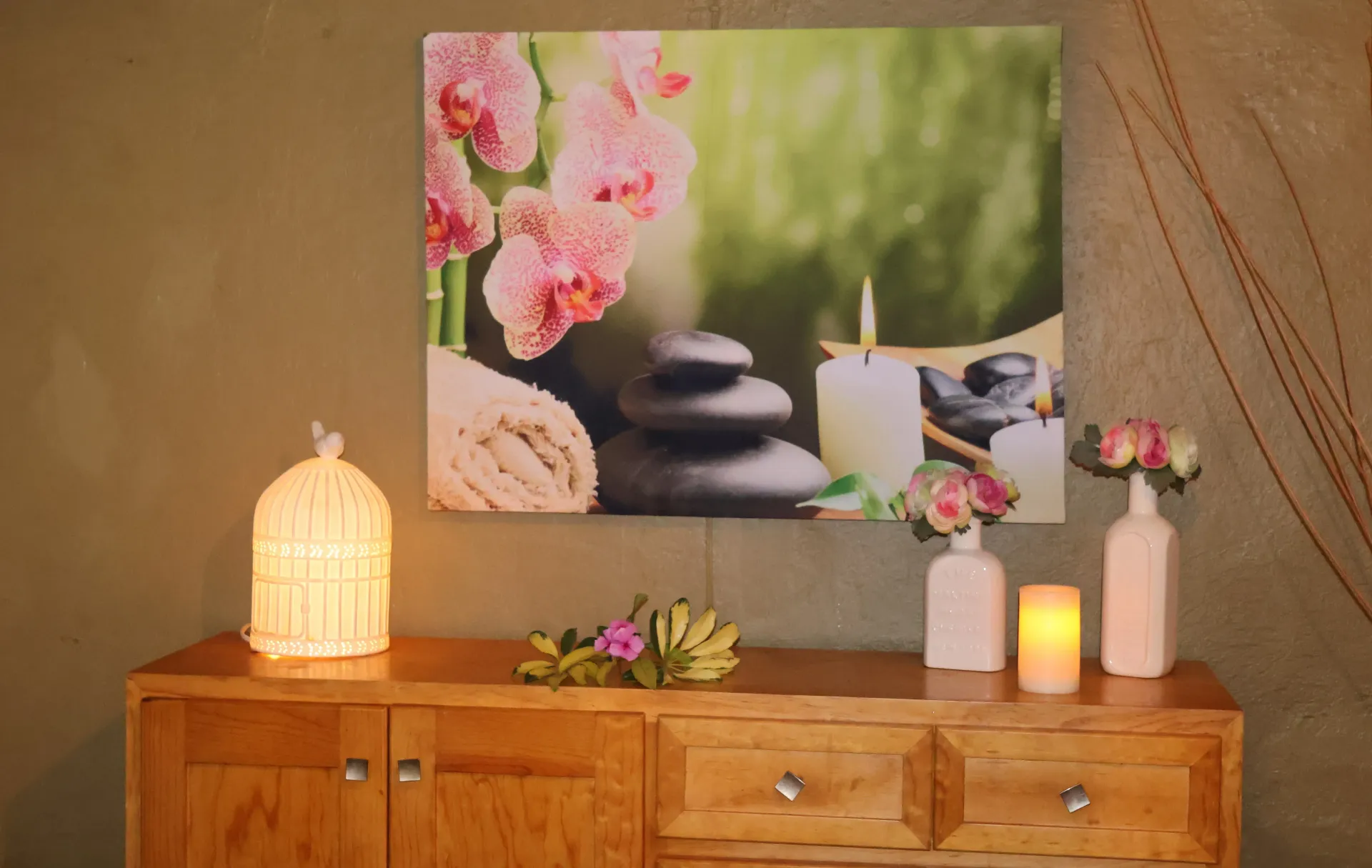 Cómoda de madera con velas encendidas, flores rosas y una lámina decorativa de pared con efecto de piedra de spa.