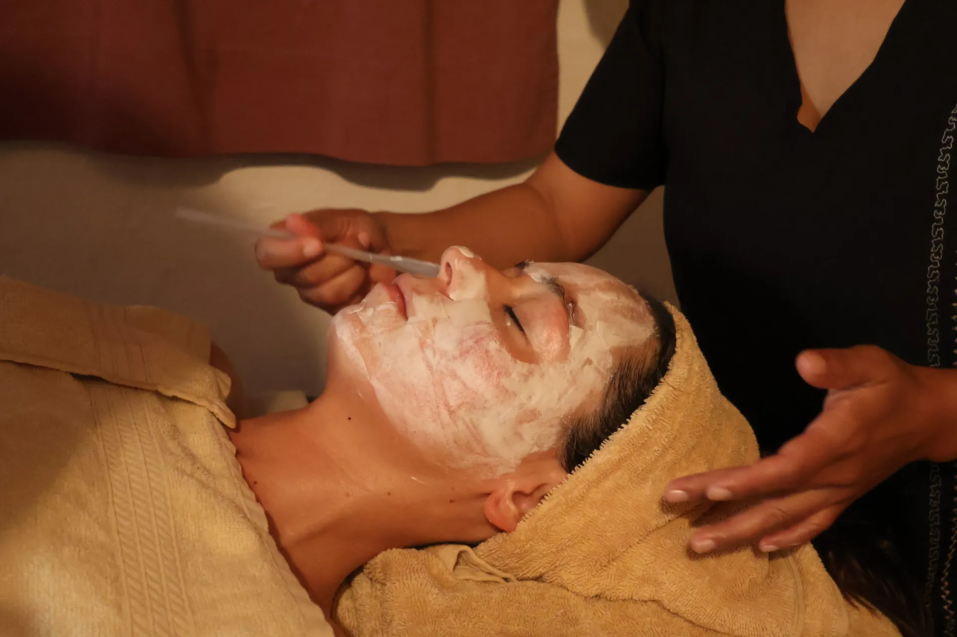 Tratamiento facial de spa con una clienta cubierta de crema mientras una esteticista aplica el producto con una brocha.