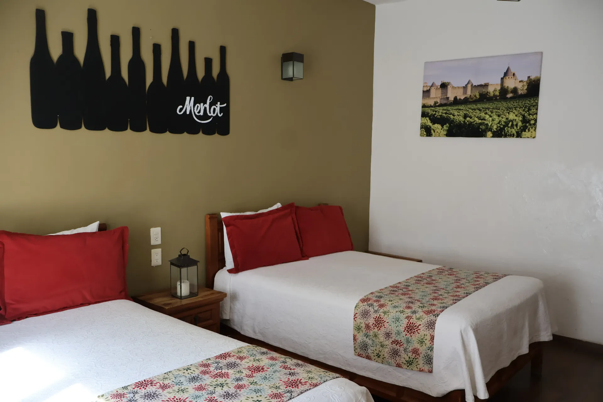 Two twin beds with red pillows in a simple hotel room, with wall art and a city photo.