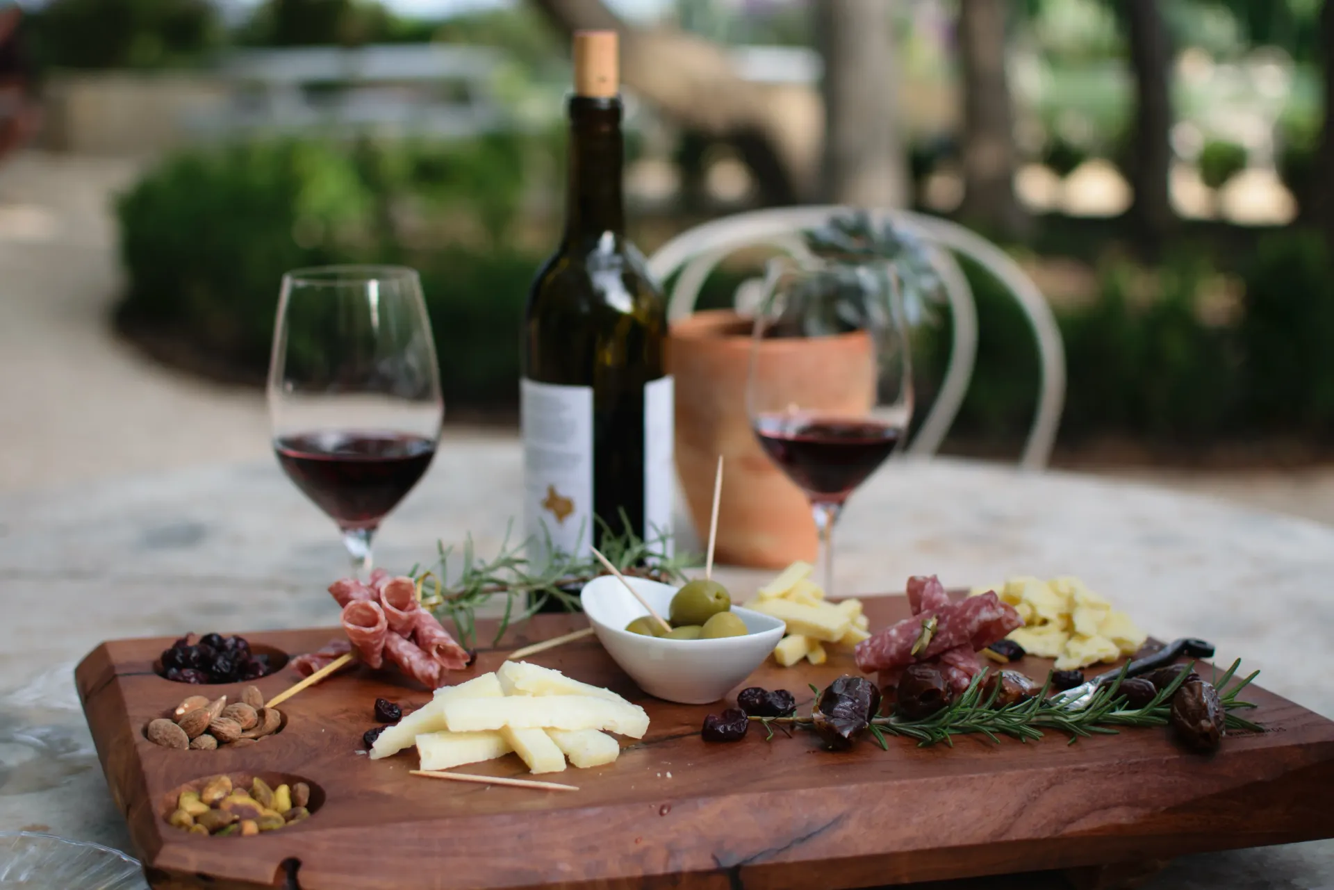 Tabla de vinos y quesos al aire libre con uvas, pan, aceitunas y dos copas de vino tinto.