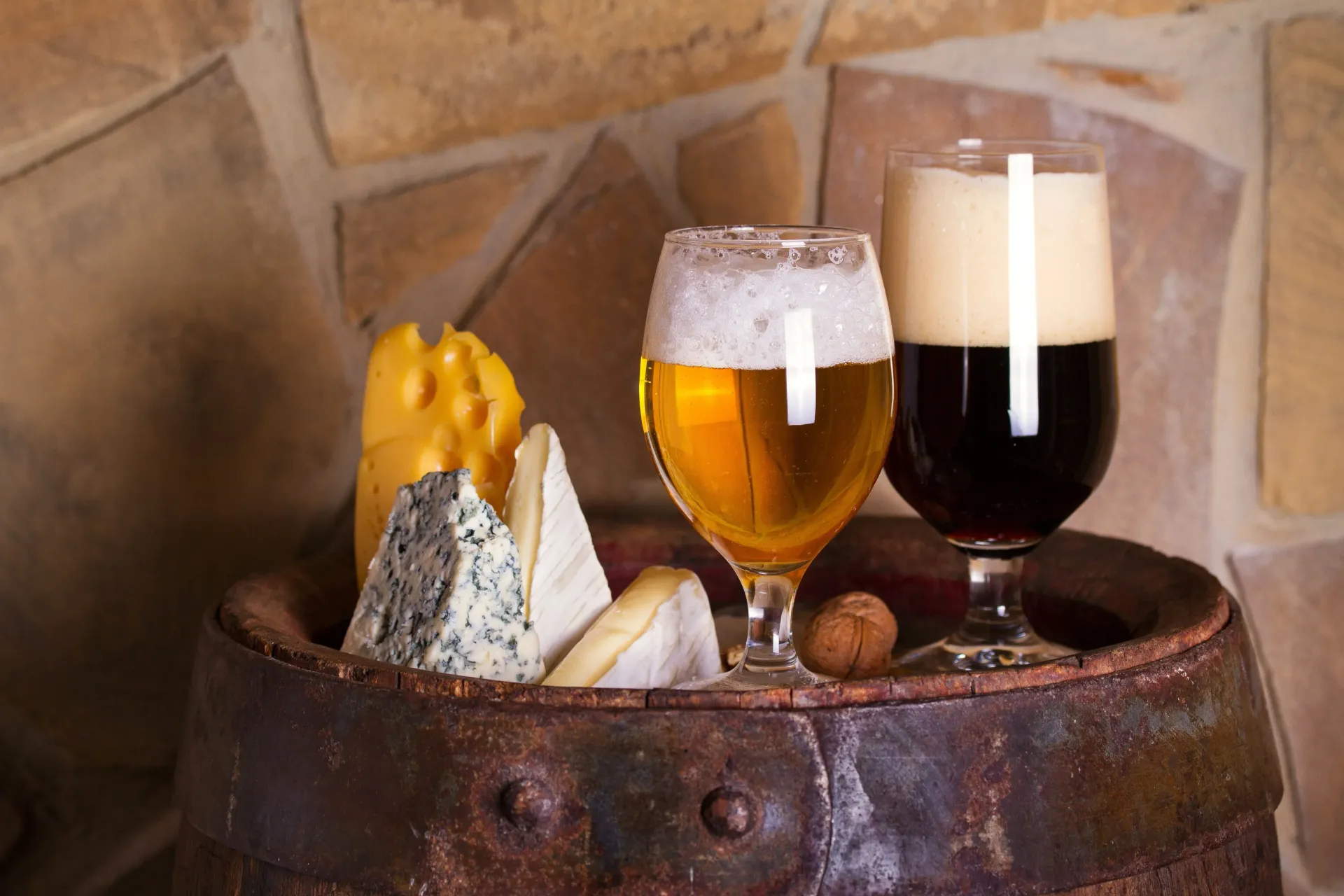 Dos vasos de cerveza espumosa sobre un barril rústico con queso y flores amarillas.