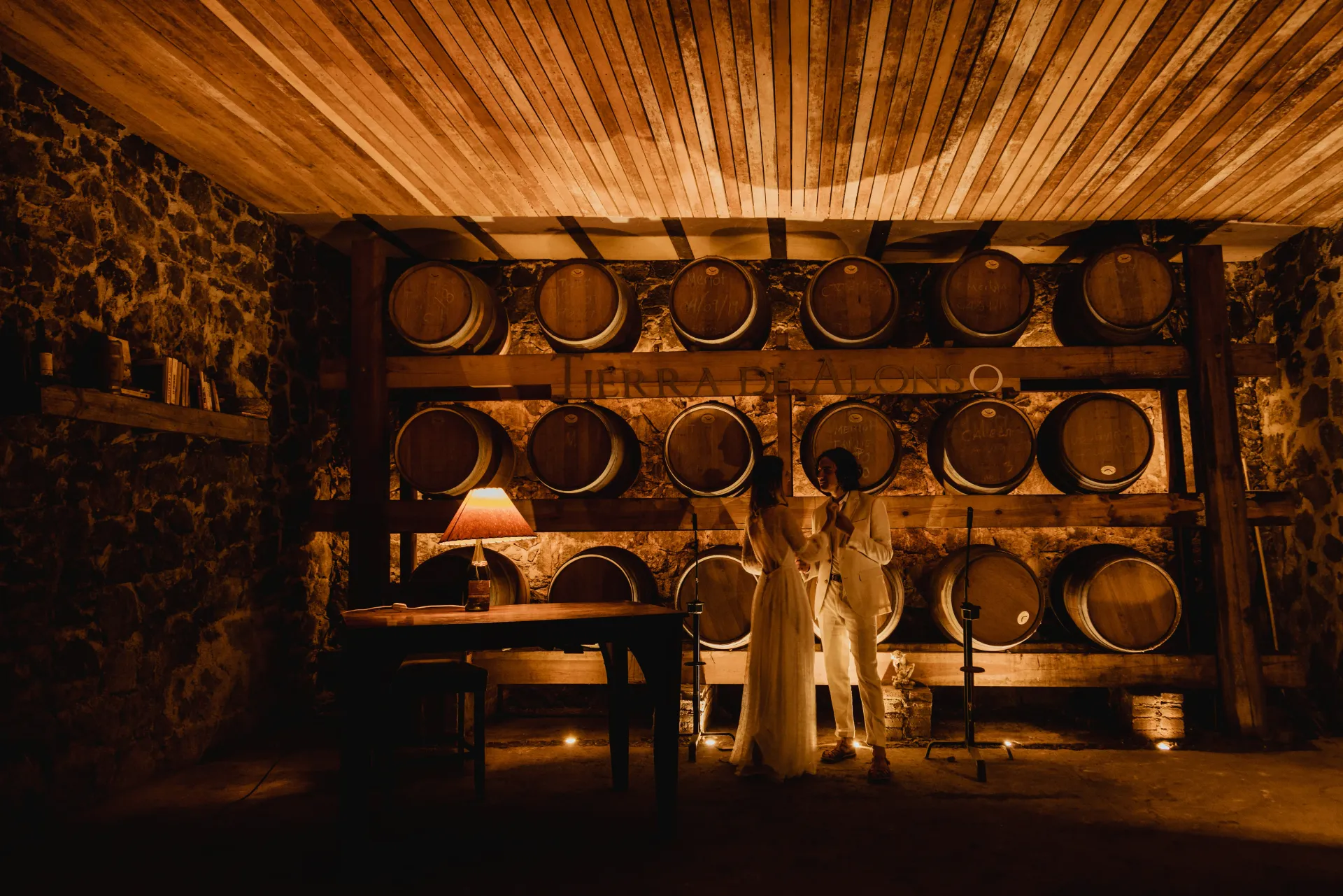 Bodega con iluminación cálida, hileras de barricas de madera y una mesita con una lámpara.