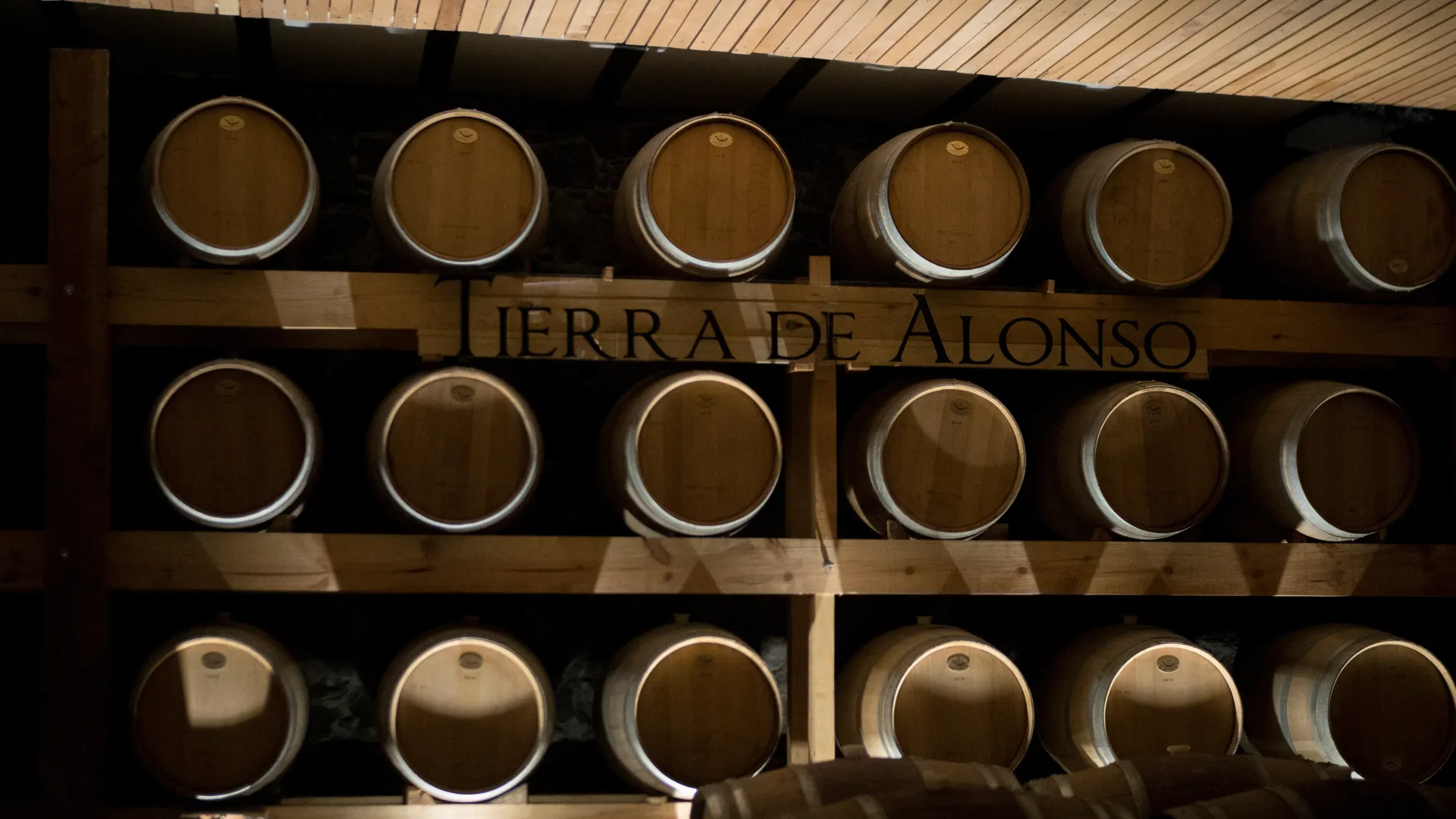 Rows of wooden barrels on shelves in a dim cellar, with “TIERRA DE ALONSO” visible in the center.