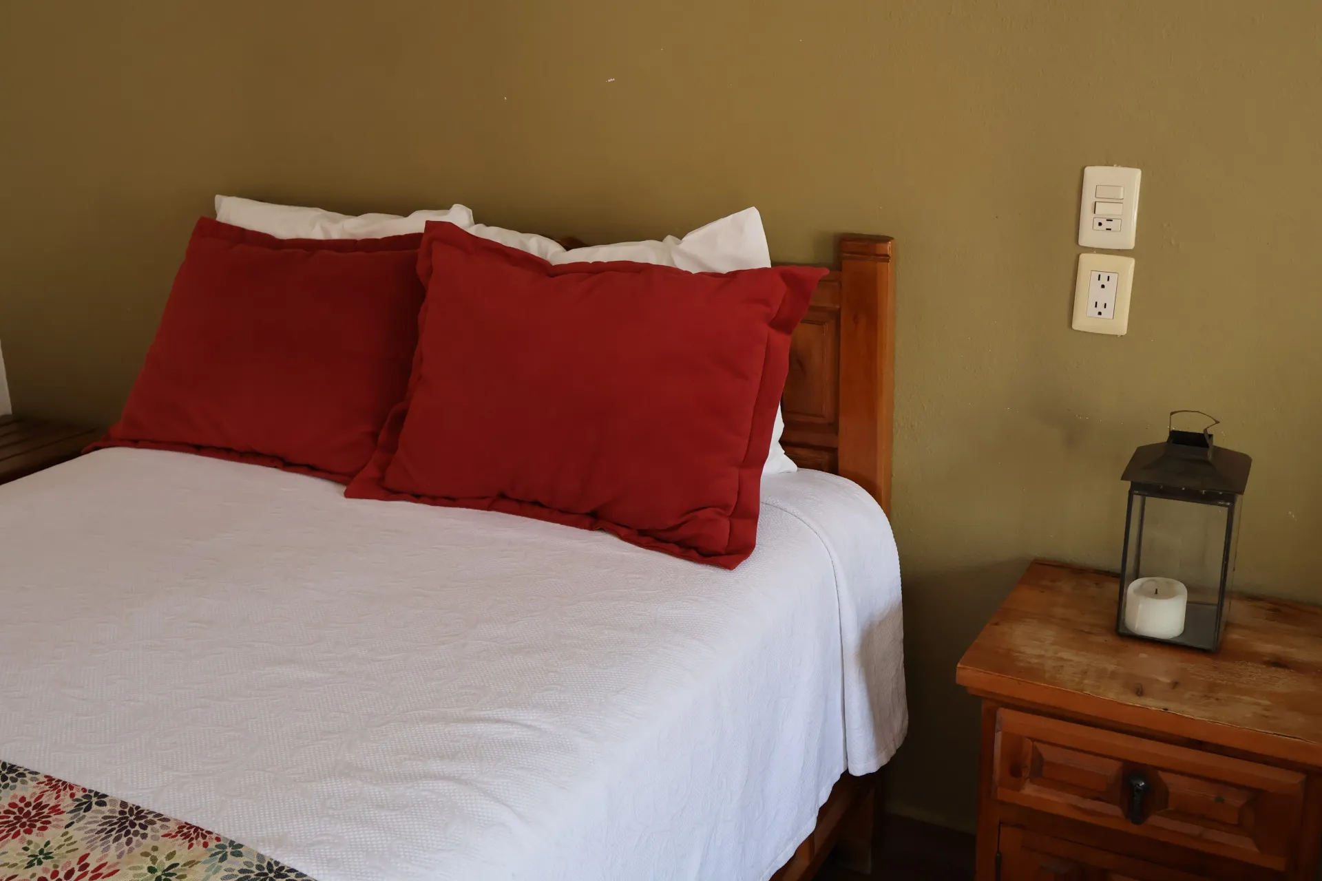 Cama con ropa de cama blanca y almohadas rojas junto a una mesita de noche de madera con una linterna.