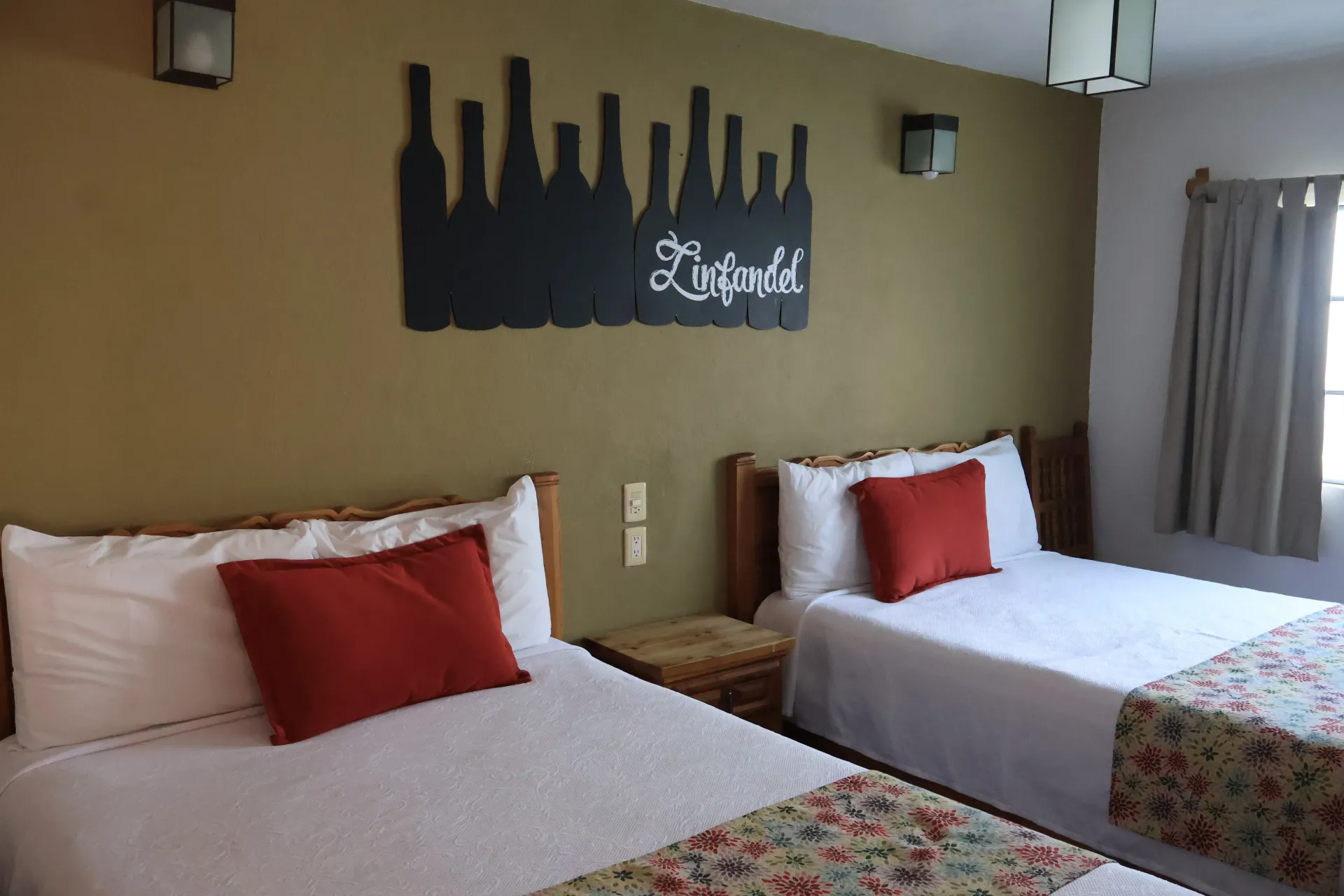 Hotel room with two white beds, red pillows, floral bedspreads, and a wall art city skyline.