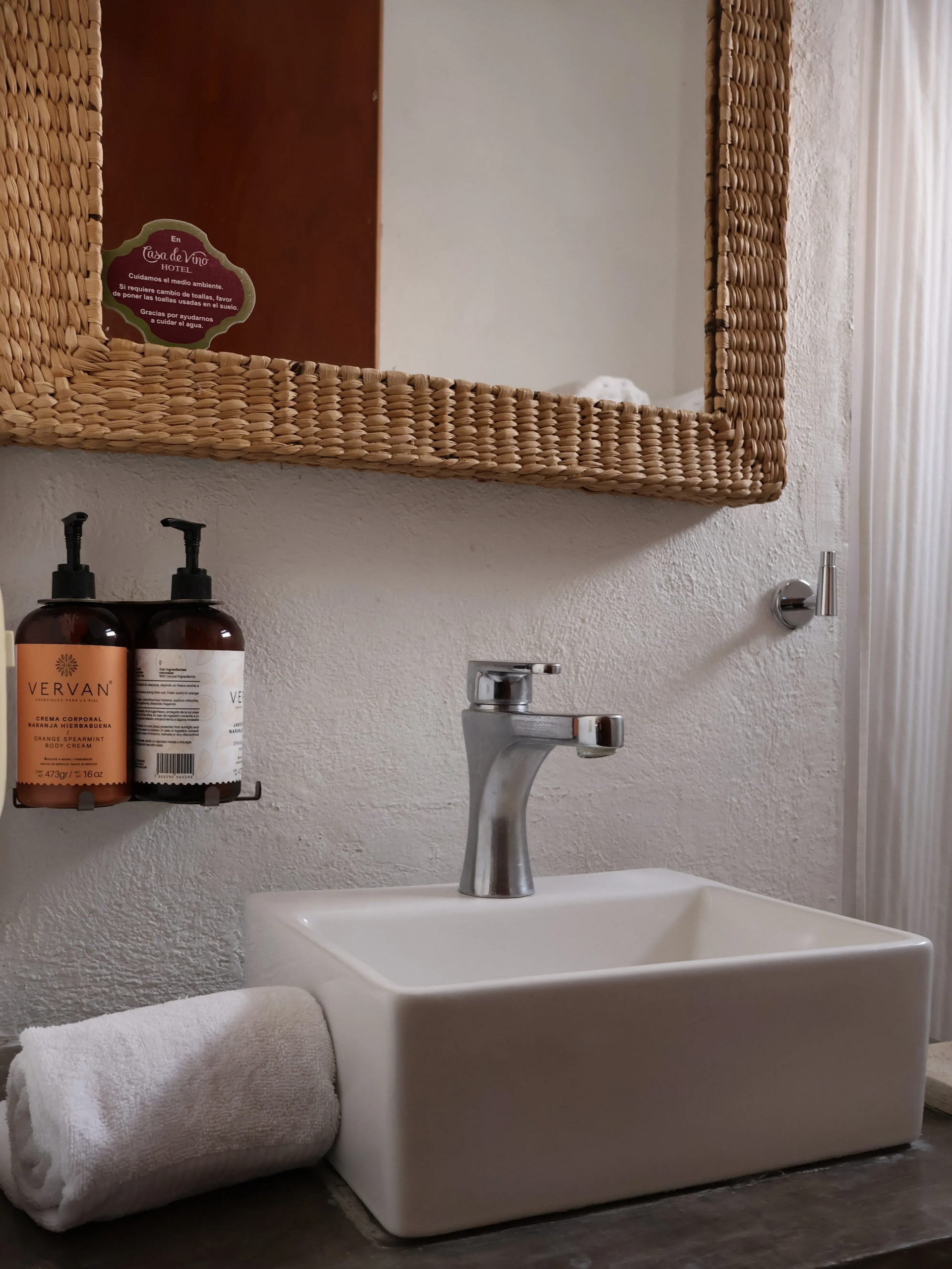 Lavabo moderno con grifo cromado, dispensadores de jabón, toalla enrollable y espejo con marco tejido.