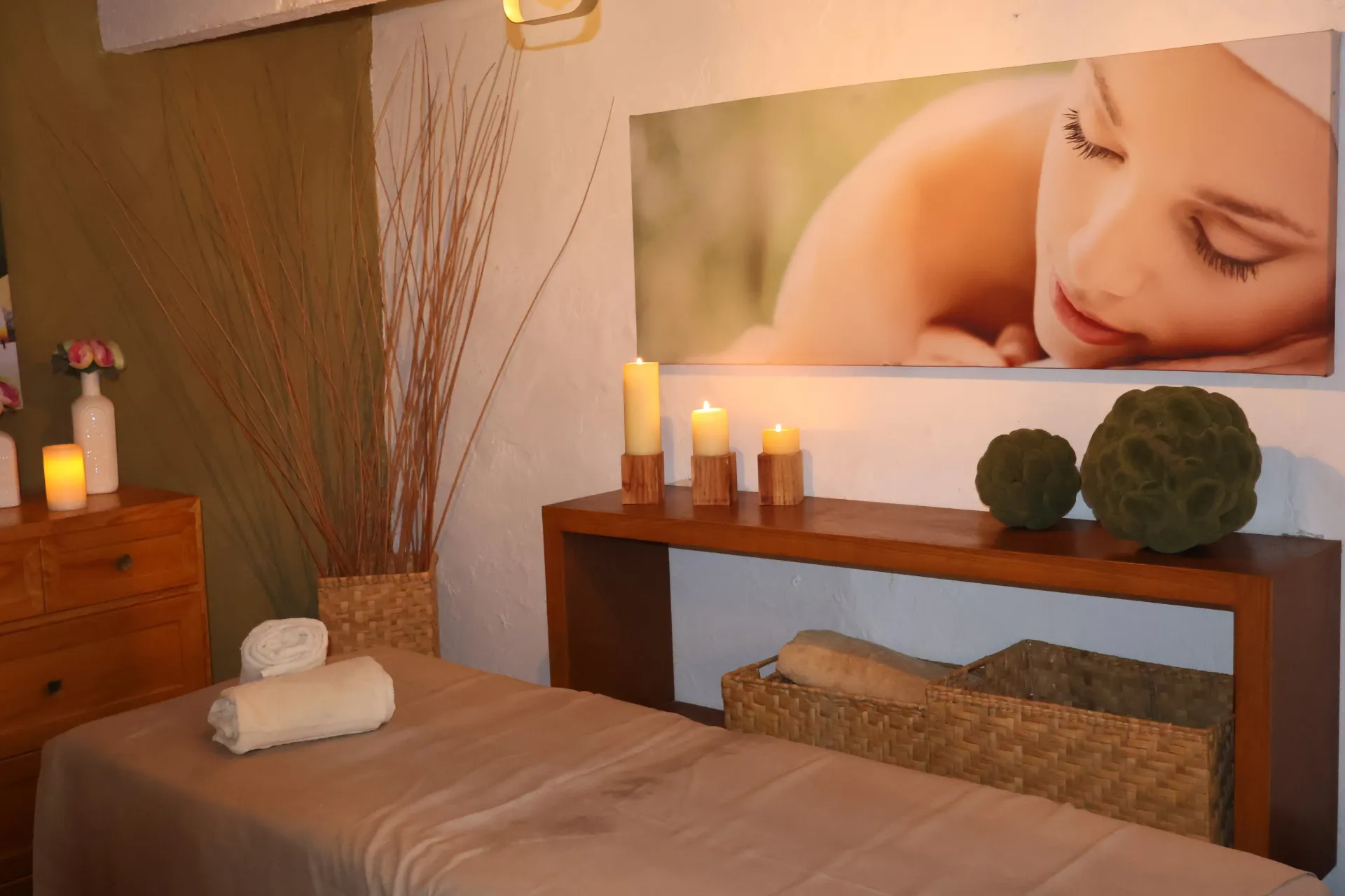 Sala de tratamientos de spa con camilla de masaje, velas, toallas y una foto de una mujer descansando sobre la pared.