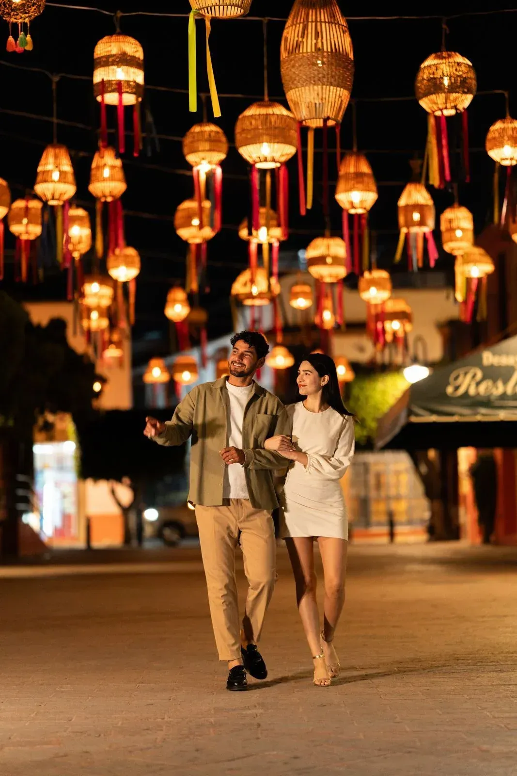 Couple walking arm in arm under glowing lanterns on a warmly lit street at night.