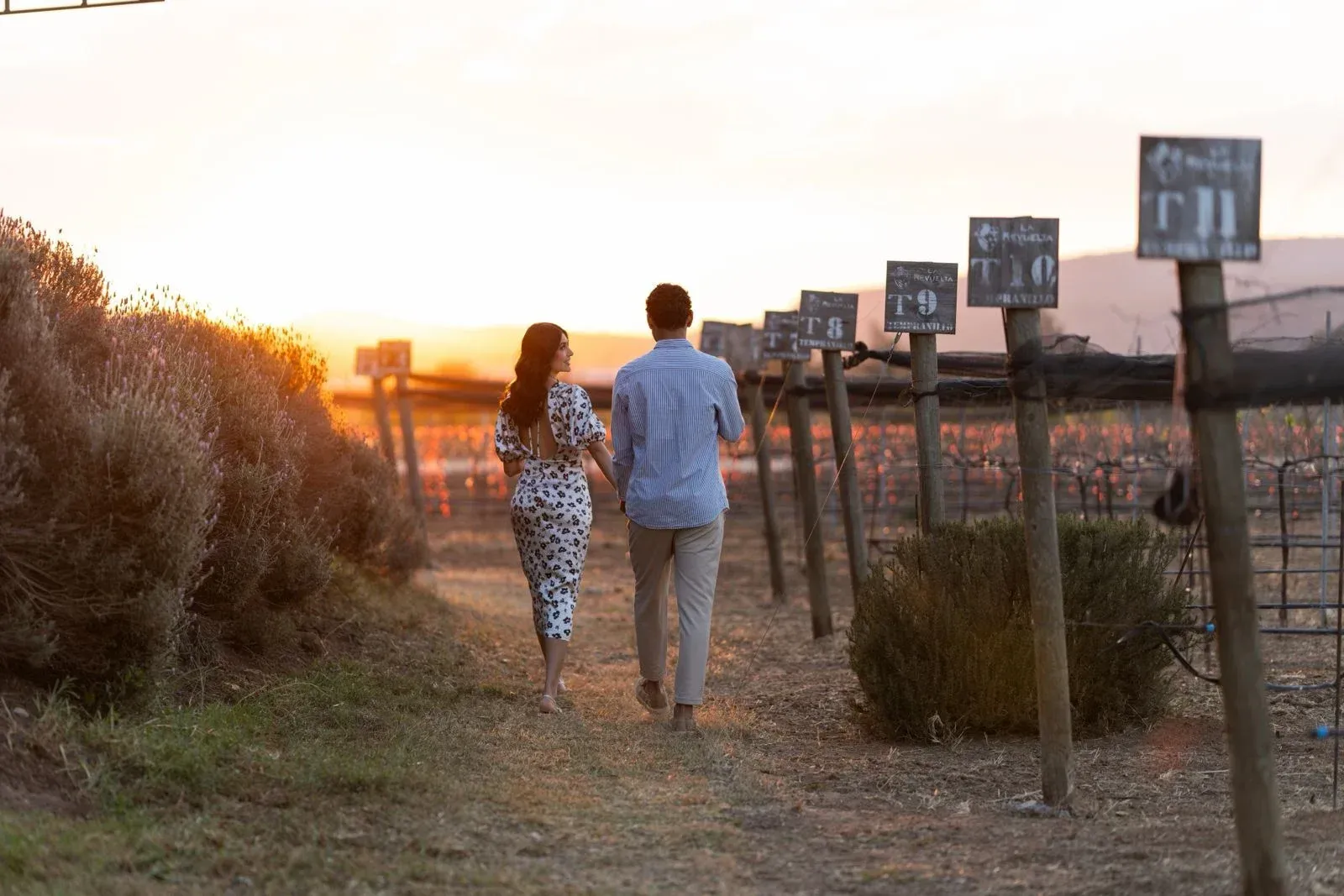 Dos personas caminan por un sendero de tierra al atardecer, junto a postes de madera en un campo abierto.