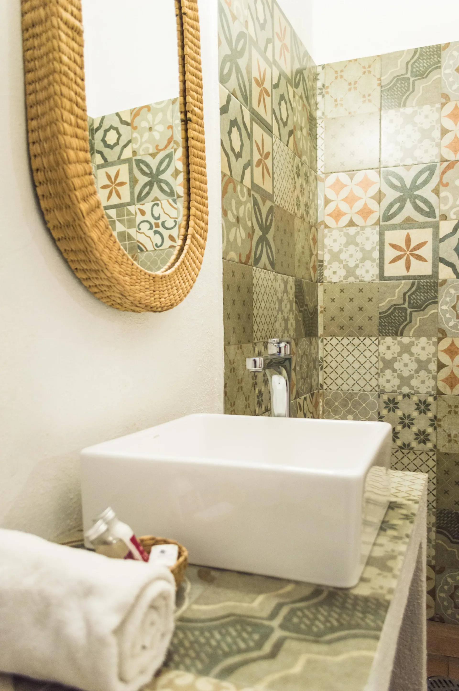Stylish bathroom with patterned tile wall, white square sink, woven mirror, and rolled towels