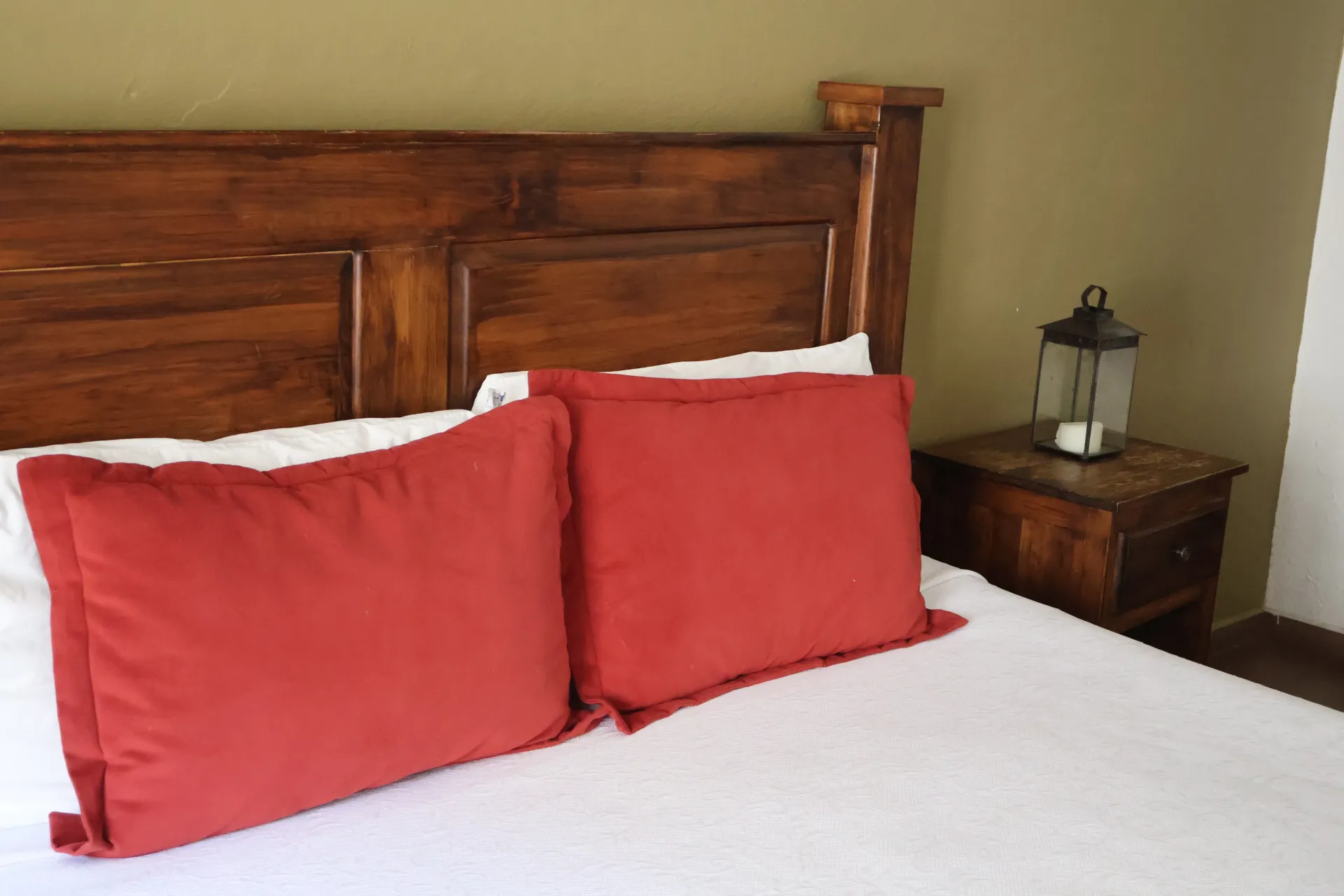 Bedroom with wooden headboard, white bedspread, two red pillows, and a bedside table with a lamp.