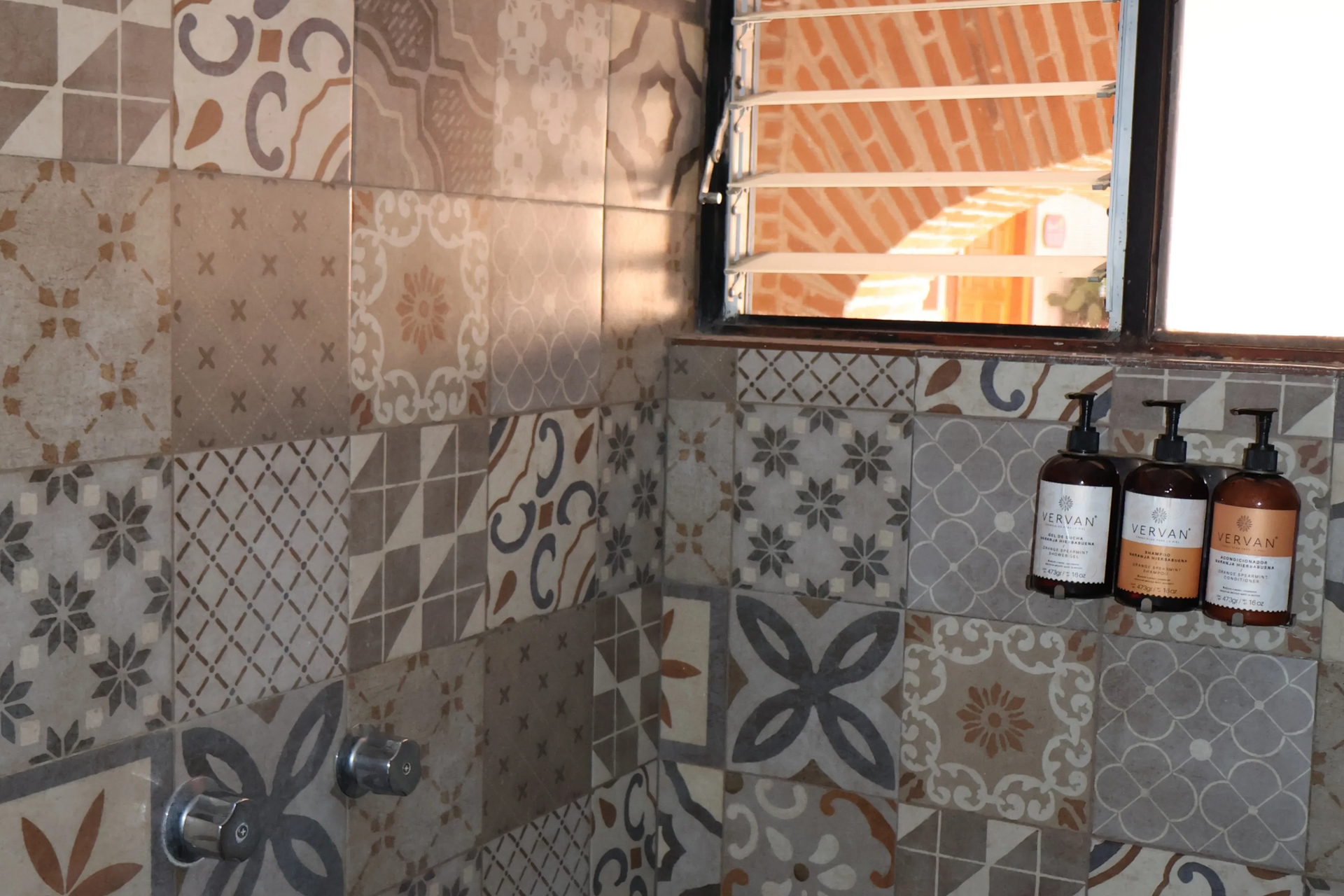 Patterned tile bathroom wall with a shower head, window, and soap bottles on a shelf