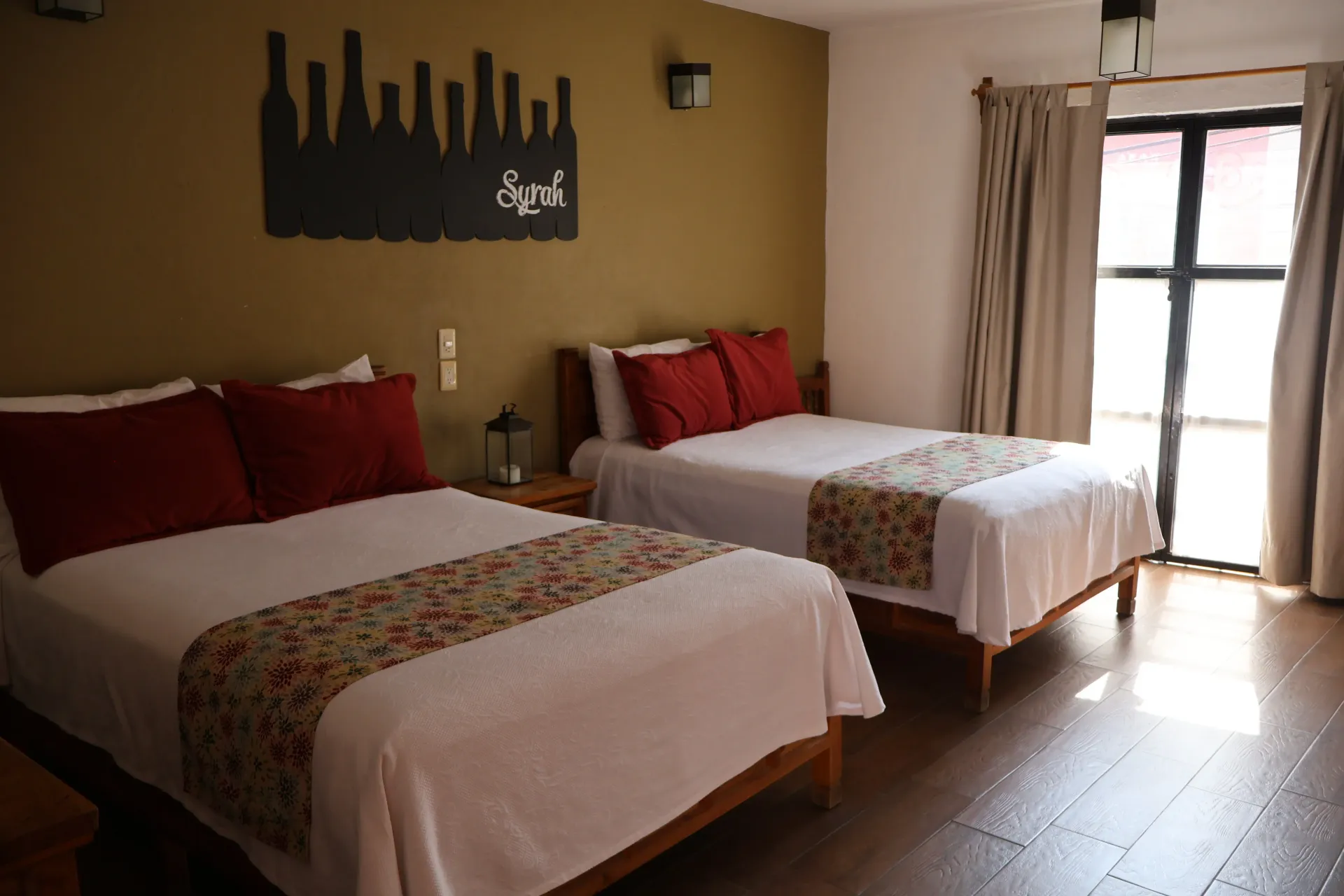 Hotel room with two neatly made beds, red pillows, beige walls, and sunlight by the window.
