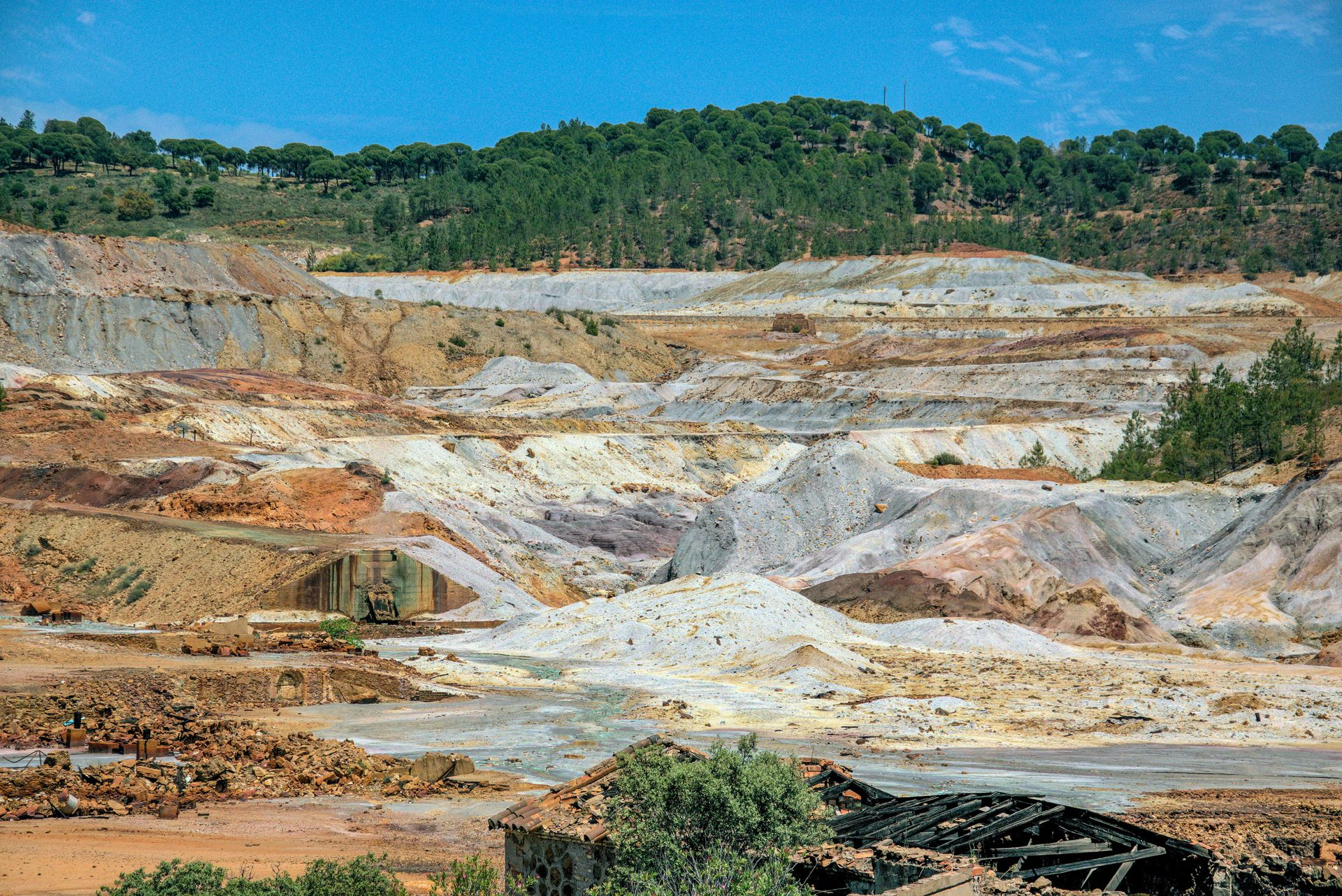 Mina a cielo abierto con terrazas de tierra en tonos grises, naranjas y blancos bajo una ladera cubierta de árboles verdes.