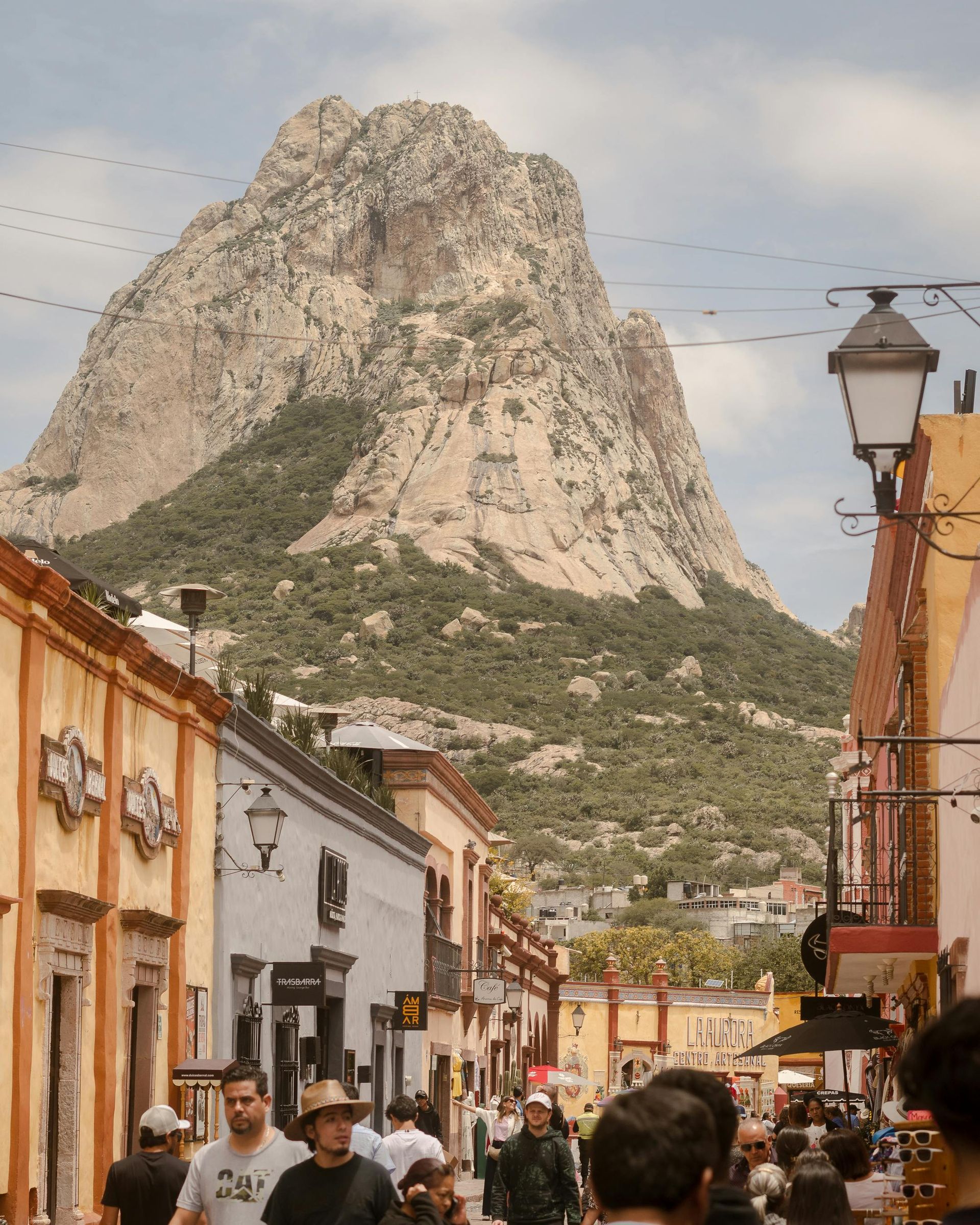 Crowded street with colorful buildings leading to a large rocky mountain under a cloudy sky