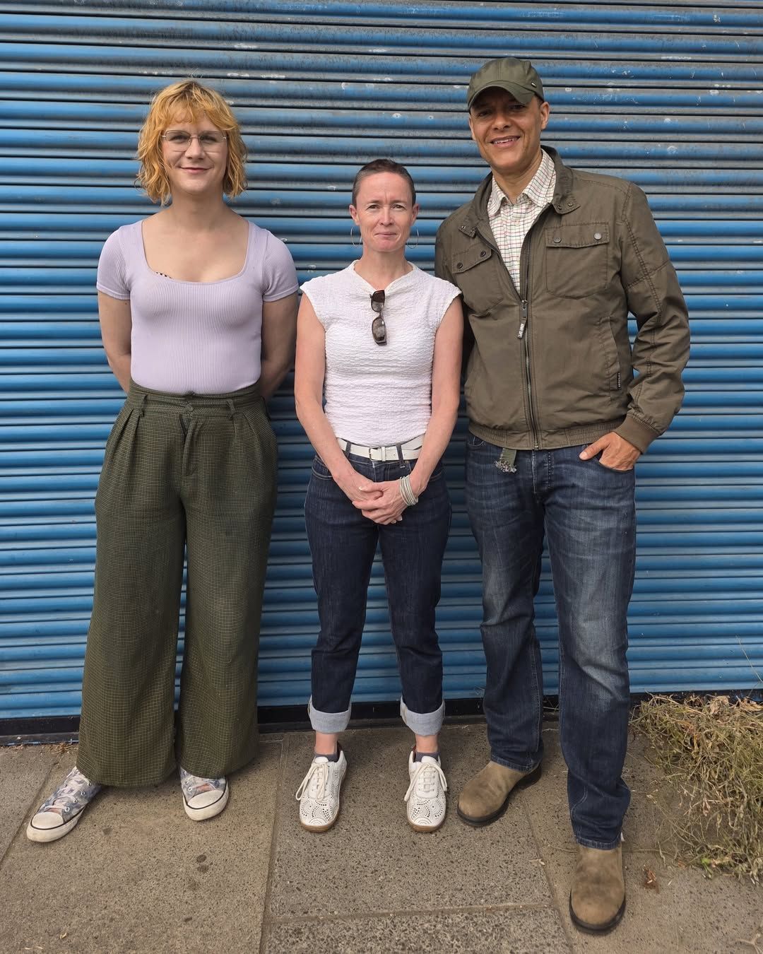 Three people standing together in front of a blue metal shutter, posing outdoors on a sidewalk.